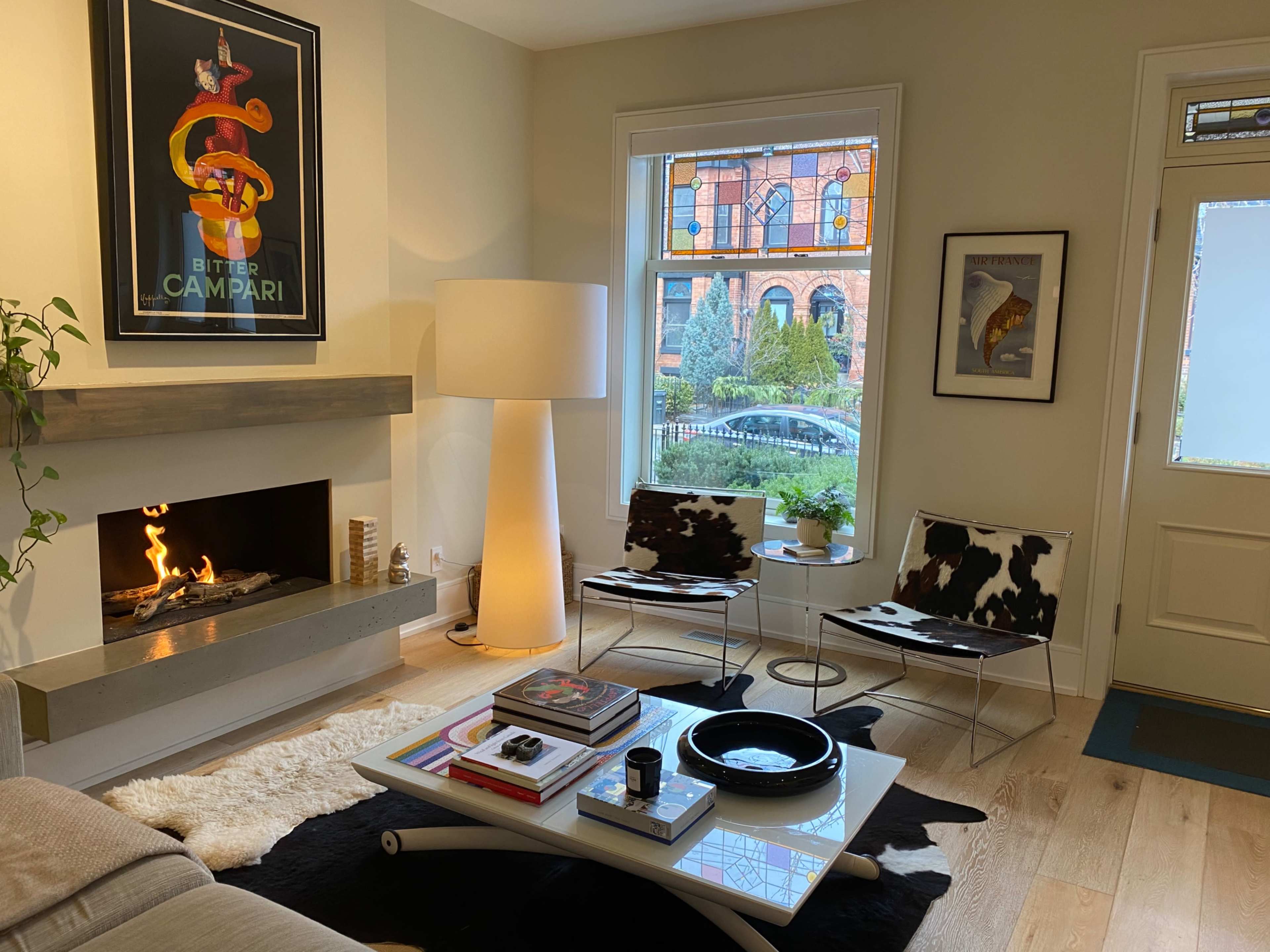 The living room features a modern fireplace, cowhide chairs, a coffee table with books, and a large lamp, with a colorful stained glass window visible outside.