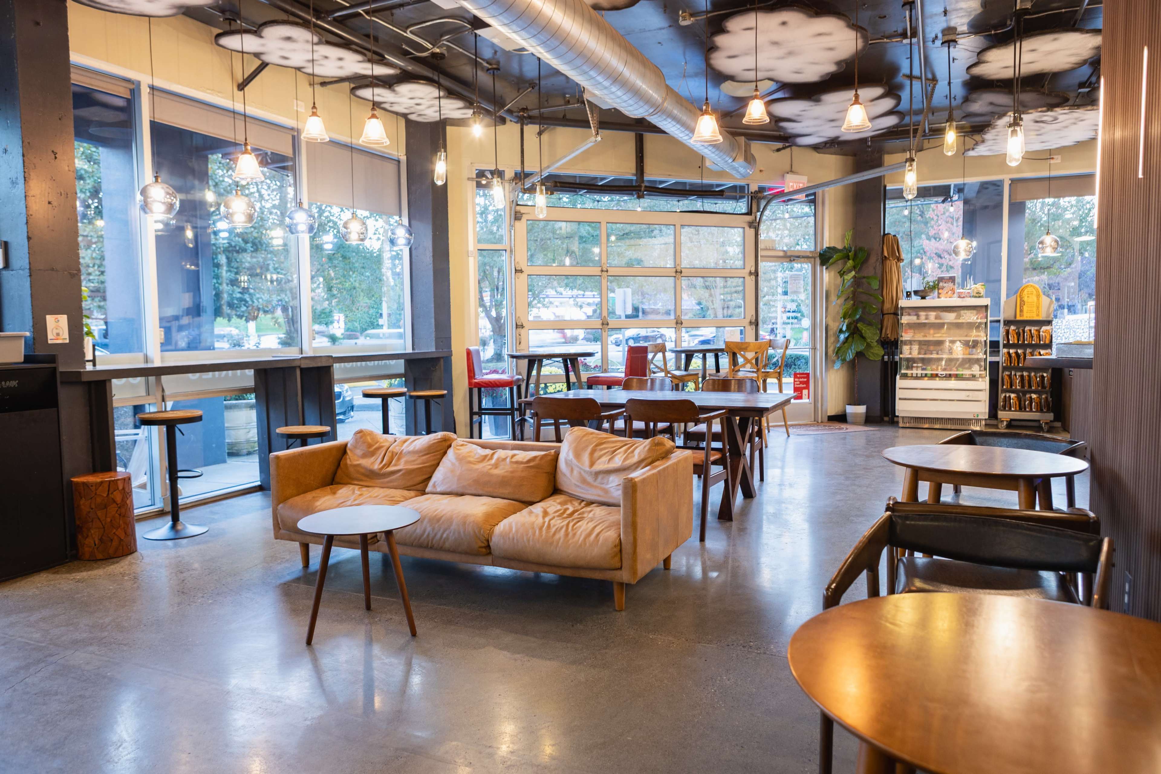 The image depicts a modern café interior featuring a mix of seating options, including a tan couch, wooden tables, and high-top bar stools, with large windows allowing natural light to fill the space.