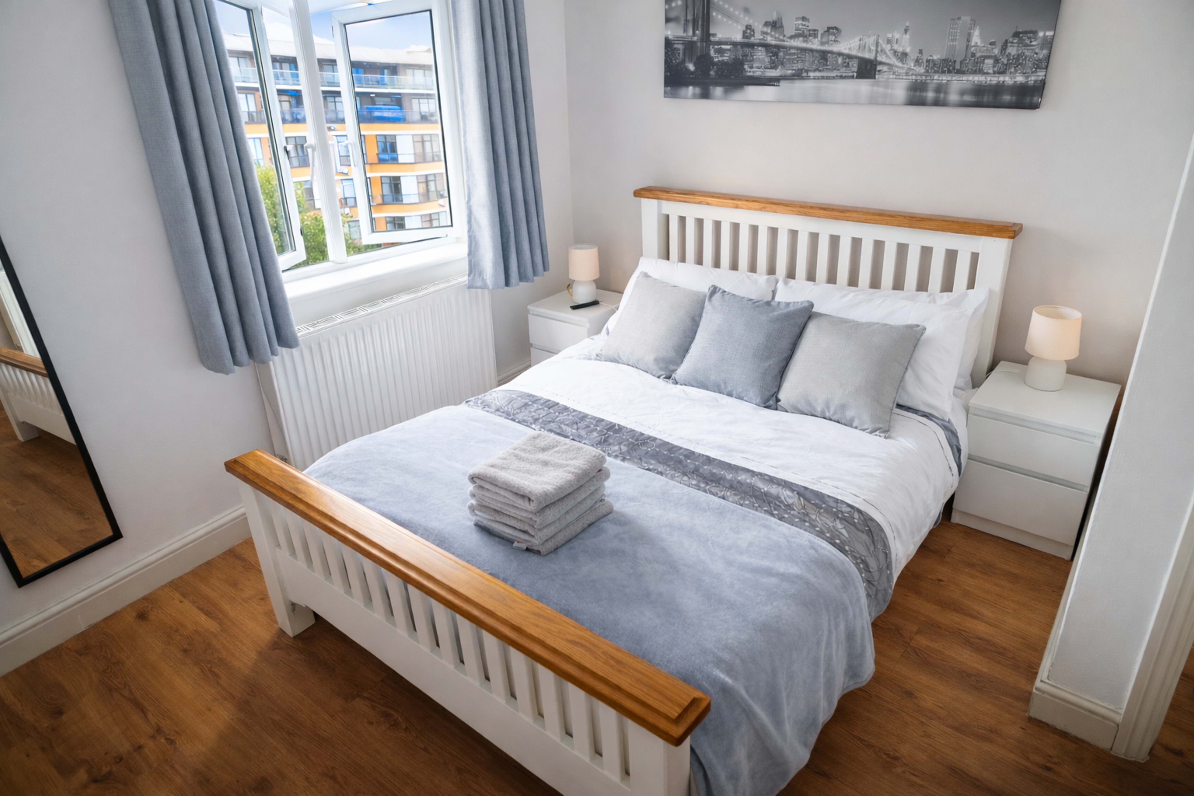 The image shows a neatly arranged bedroom with a wooden-framed bed, light gray bedding, and a window overlooking an urban landscape.