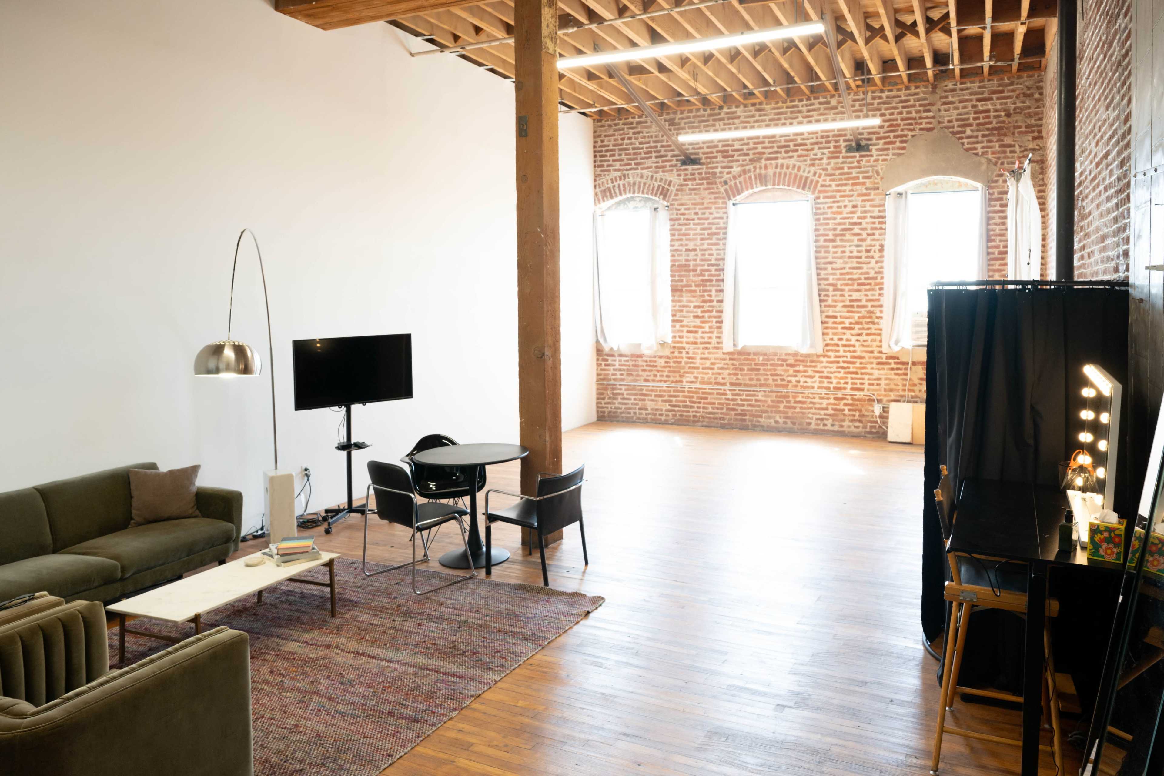 The image shows a spacious interior with brick walls, wooden floors, a sofa, a table with chairs, and large windows allowing natural light.