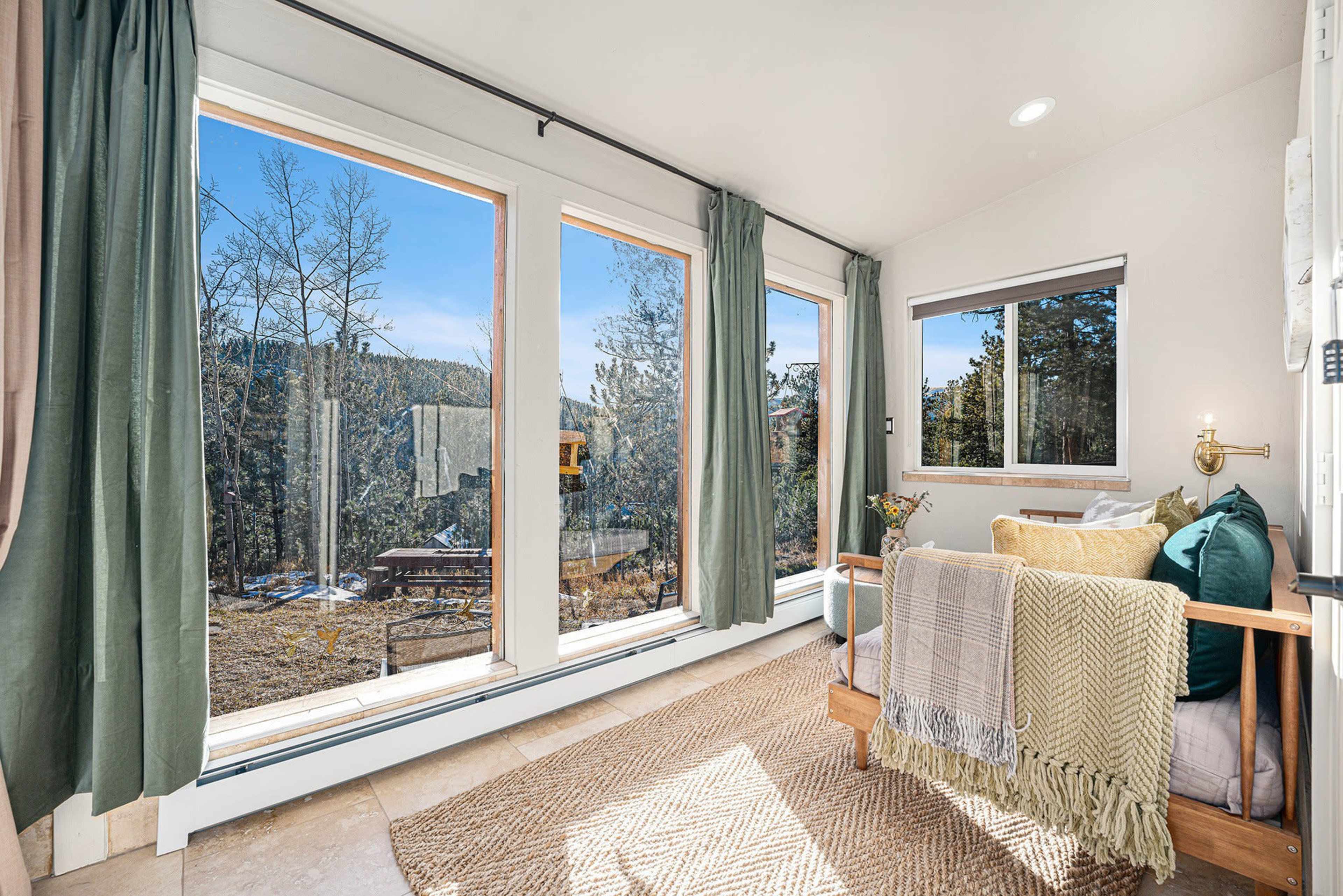 A bright room features large windows with views of trees and outdoor seating, adorned with a cozy seating area and decorative throws.