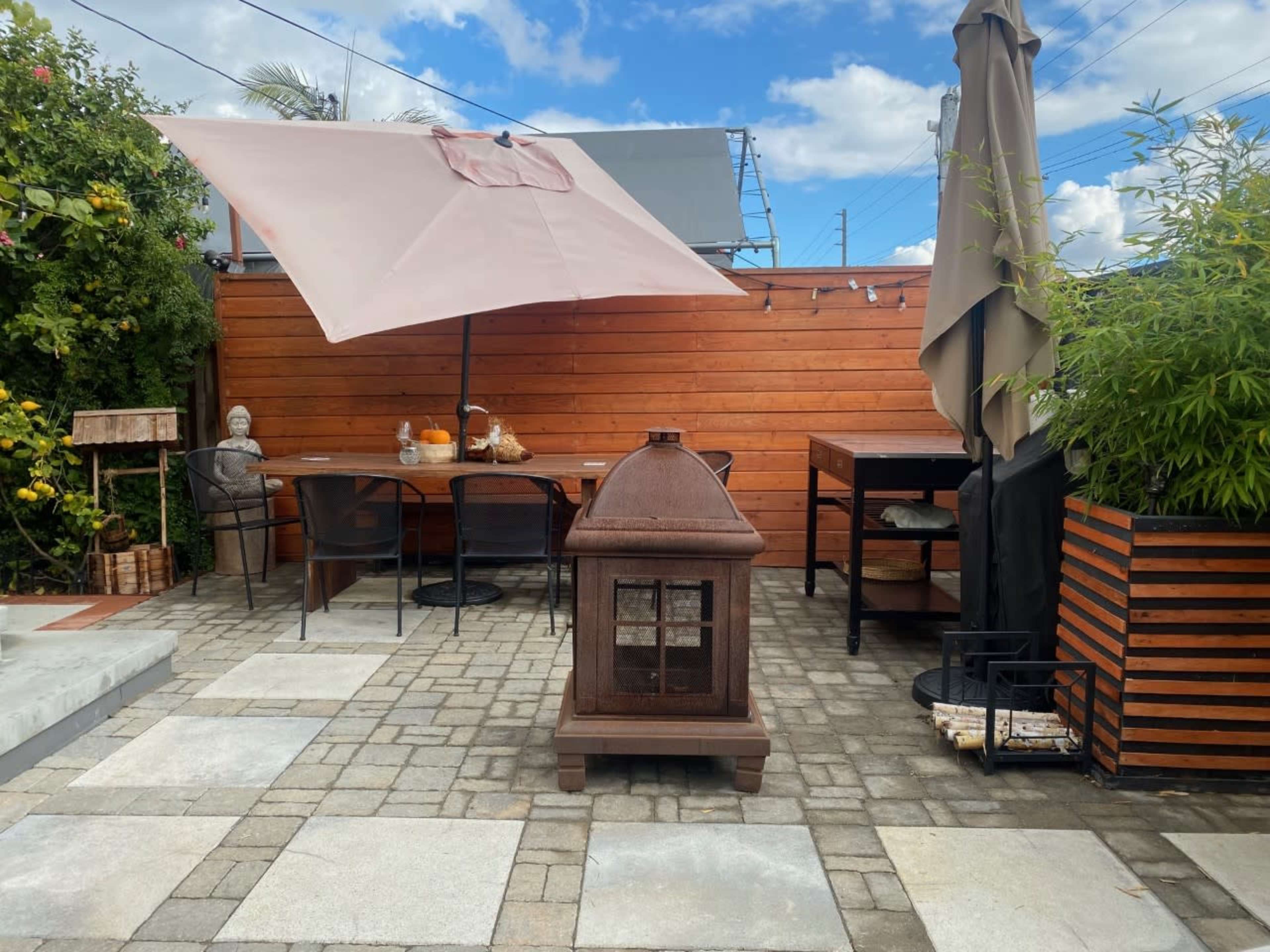 The patio features a gas fire pit, a large umbrella, a dining table, and a garden area with greenery and decorative elements.