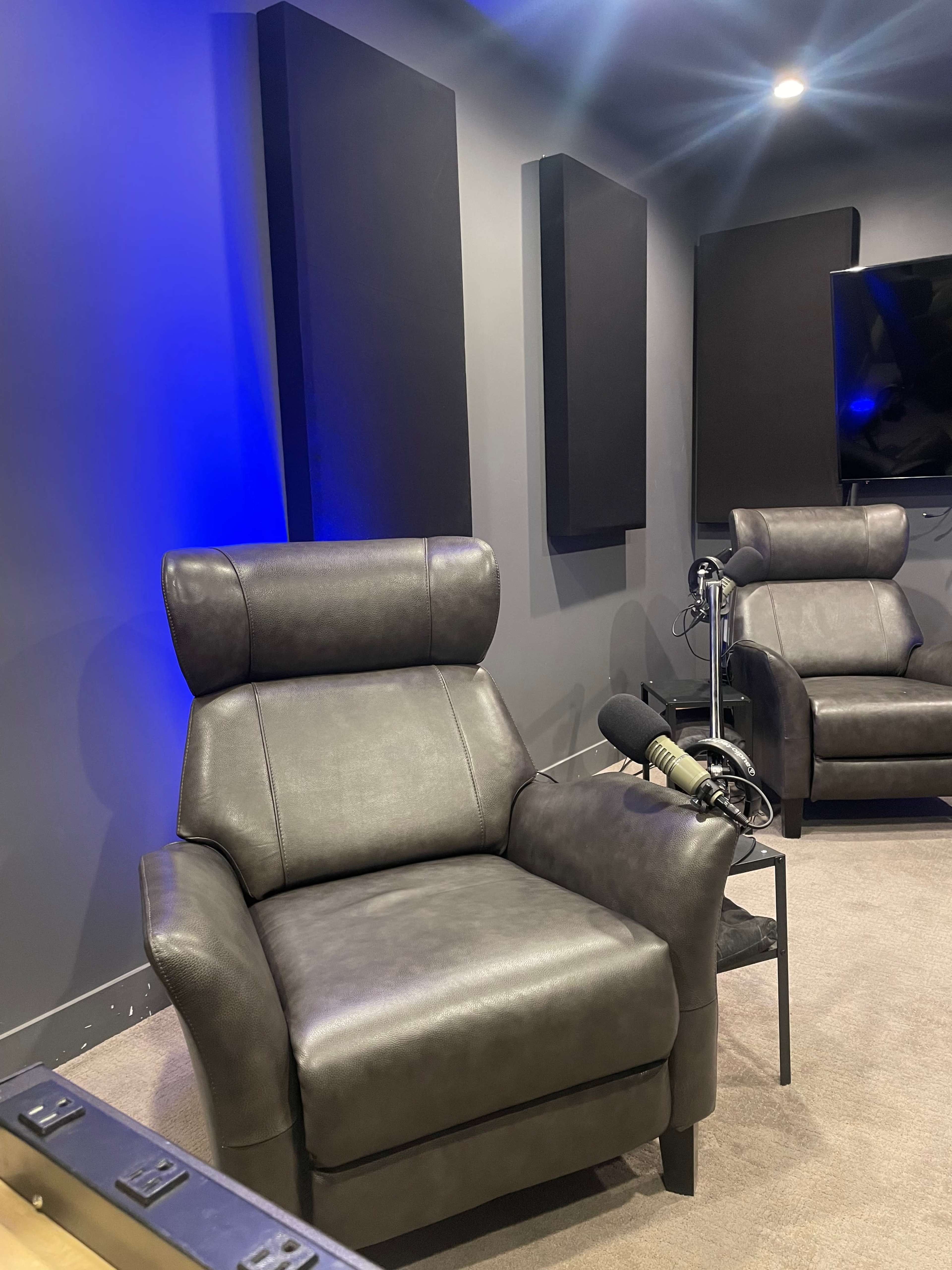 A modern recording studio setup featuring two gray leather recliner chairs positioned against a dark wall with soundproofing panels.