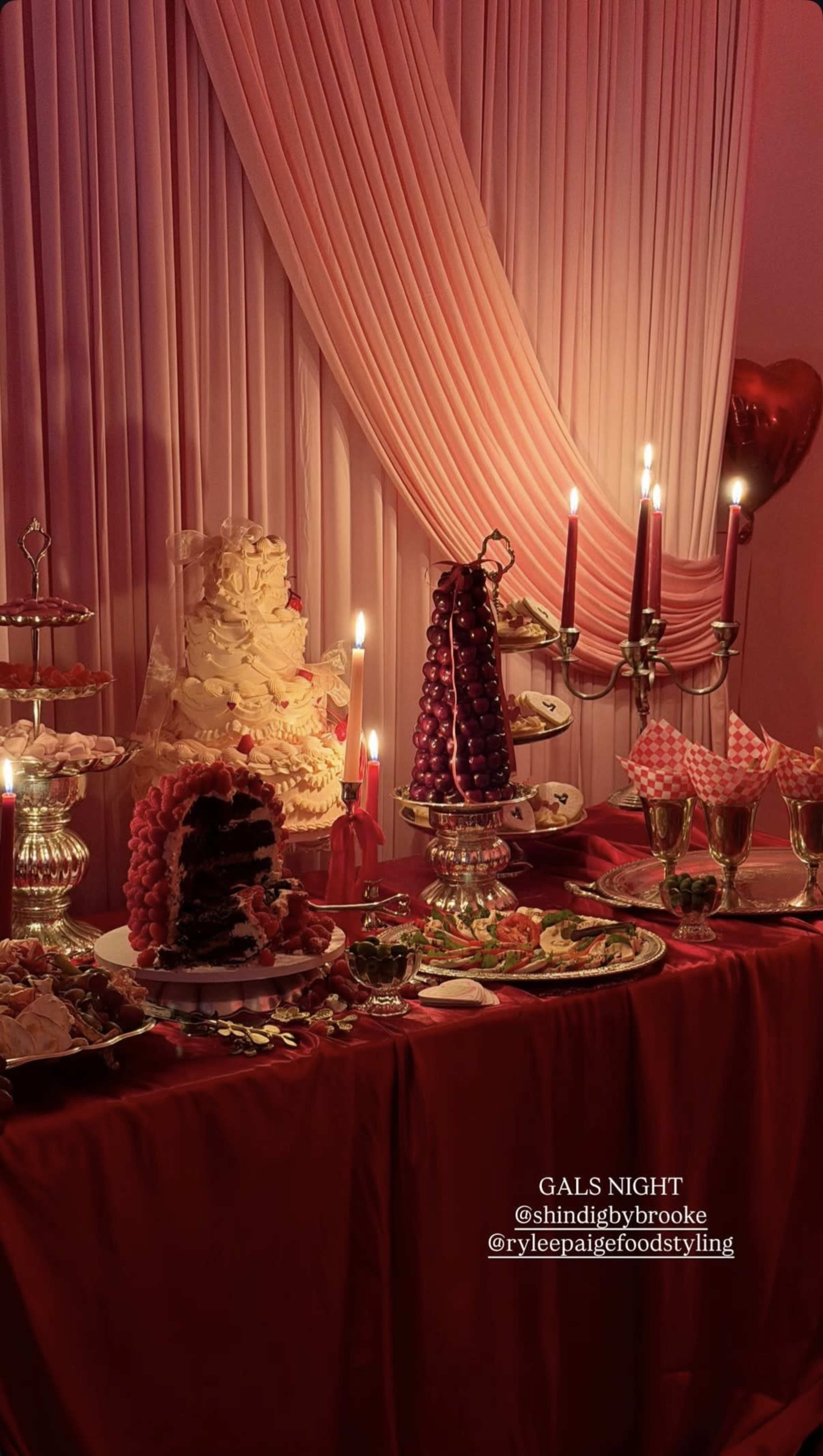 A table is set for a gathering, featuring a large cake, decorative pastries, fruit displays, and candles against a backdrop of pink drapery.