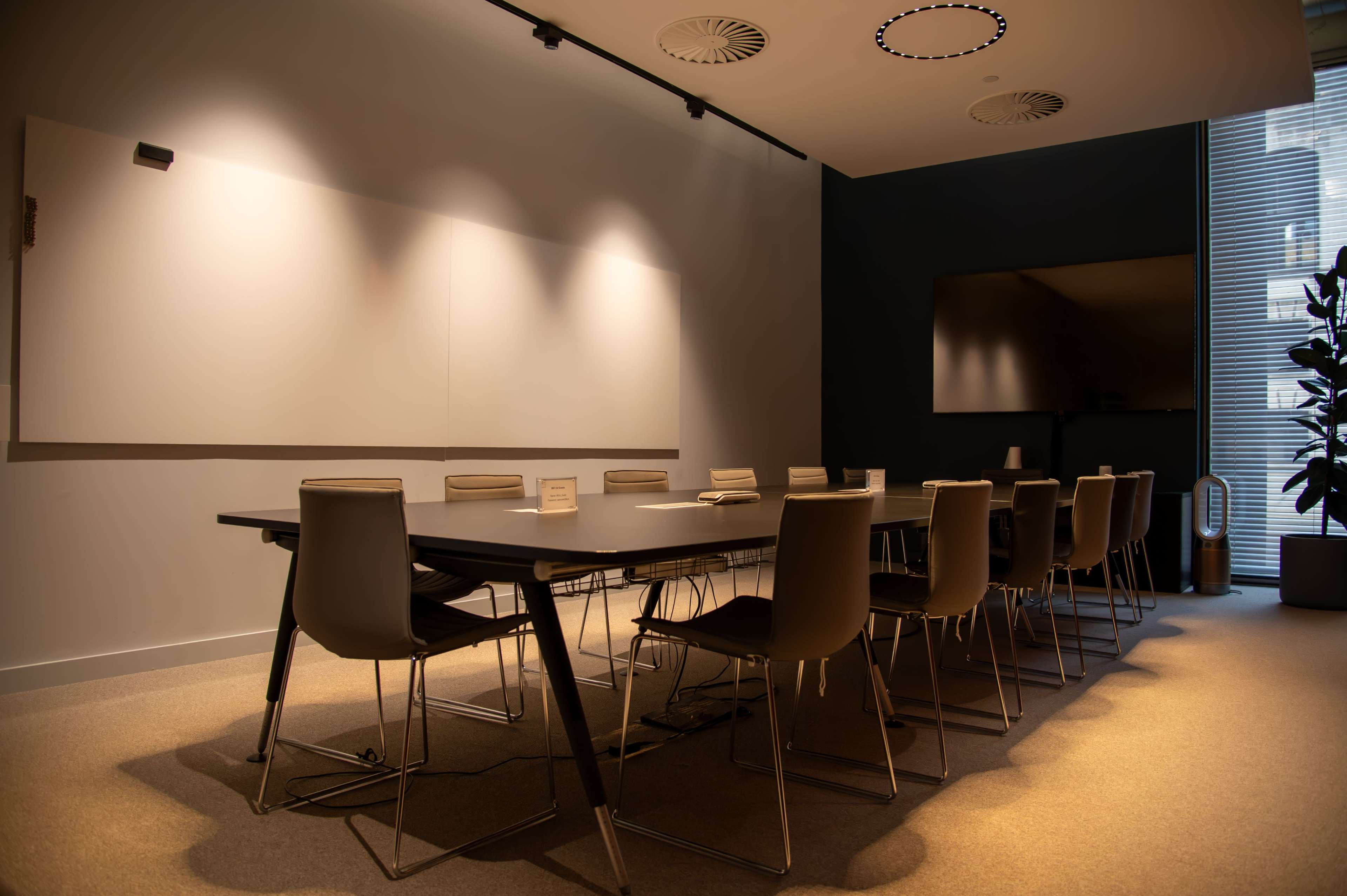 A modern conference room features a large table surrounded by multiple chairs, with a whiteboard and a screen on the walls.