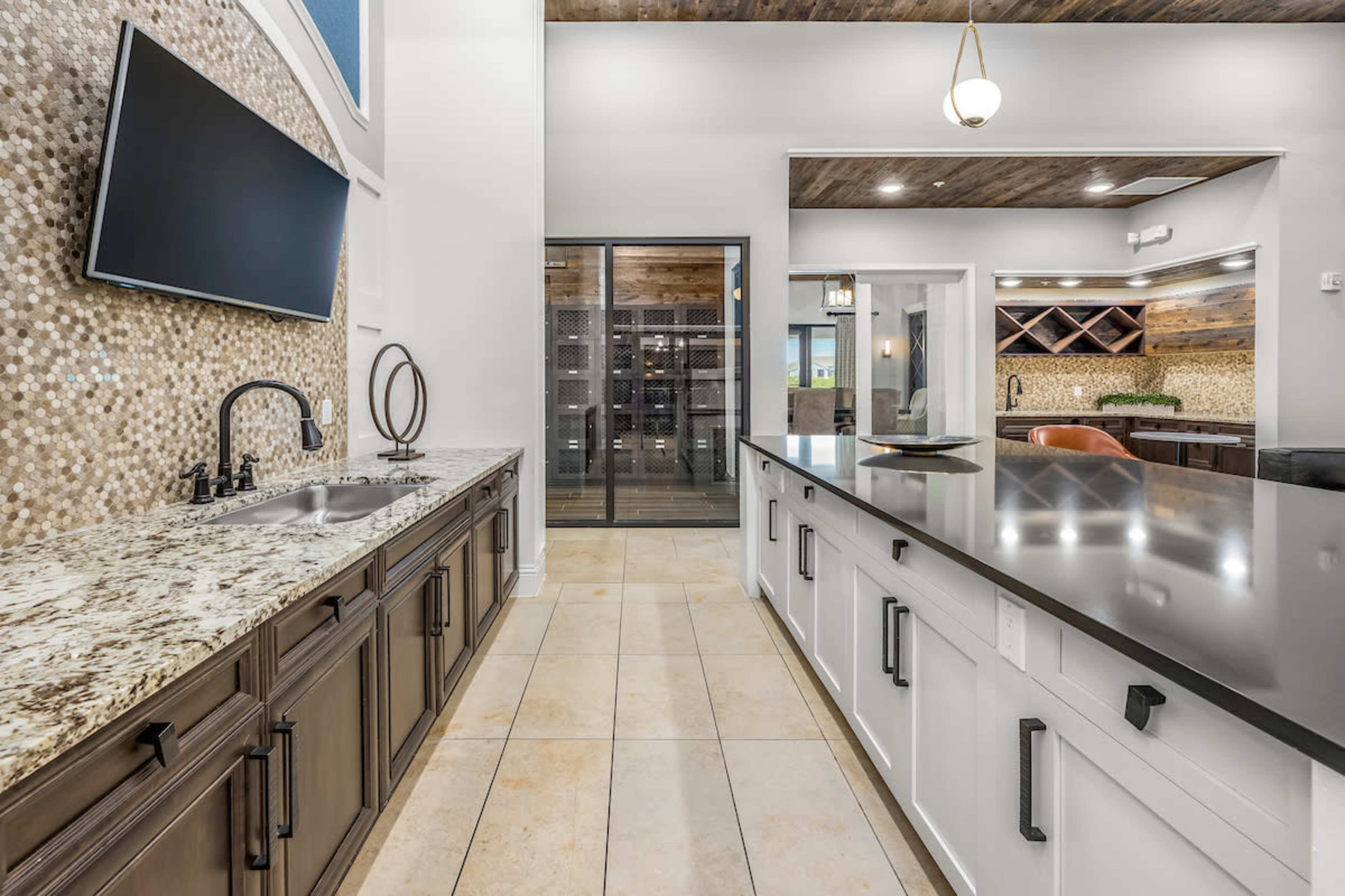 The image shows a modern kitchen with a granite countertop, a wall-mounted television, and glass doors leading to a wine cellar.