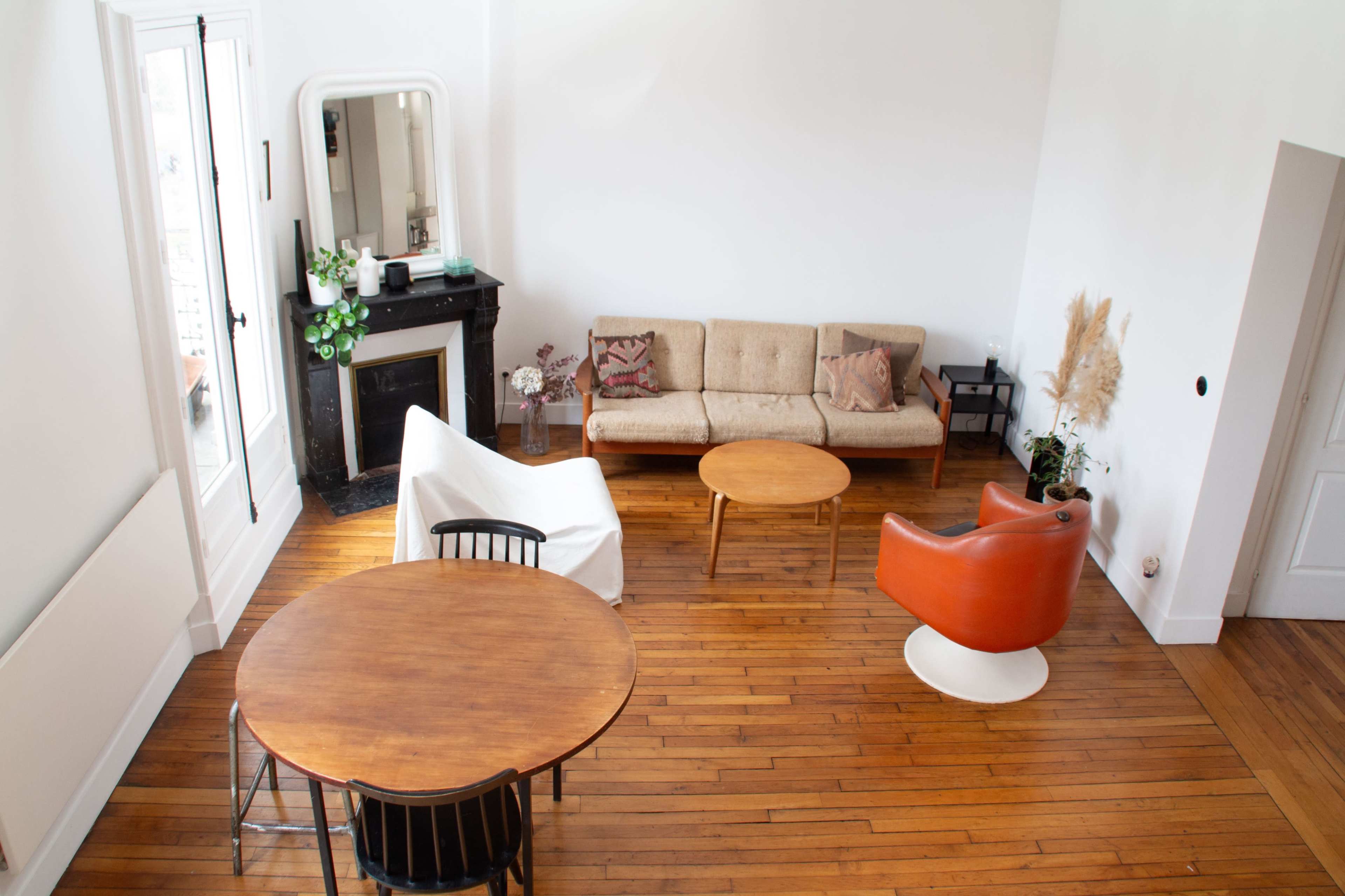 A spacious living area with a beige sofa, an orange armchair, a round wooden table, and a fireplace, all arranged on polished wooden floors.