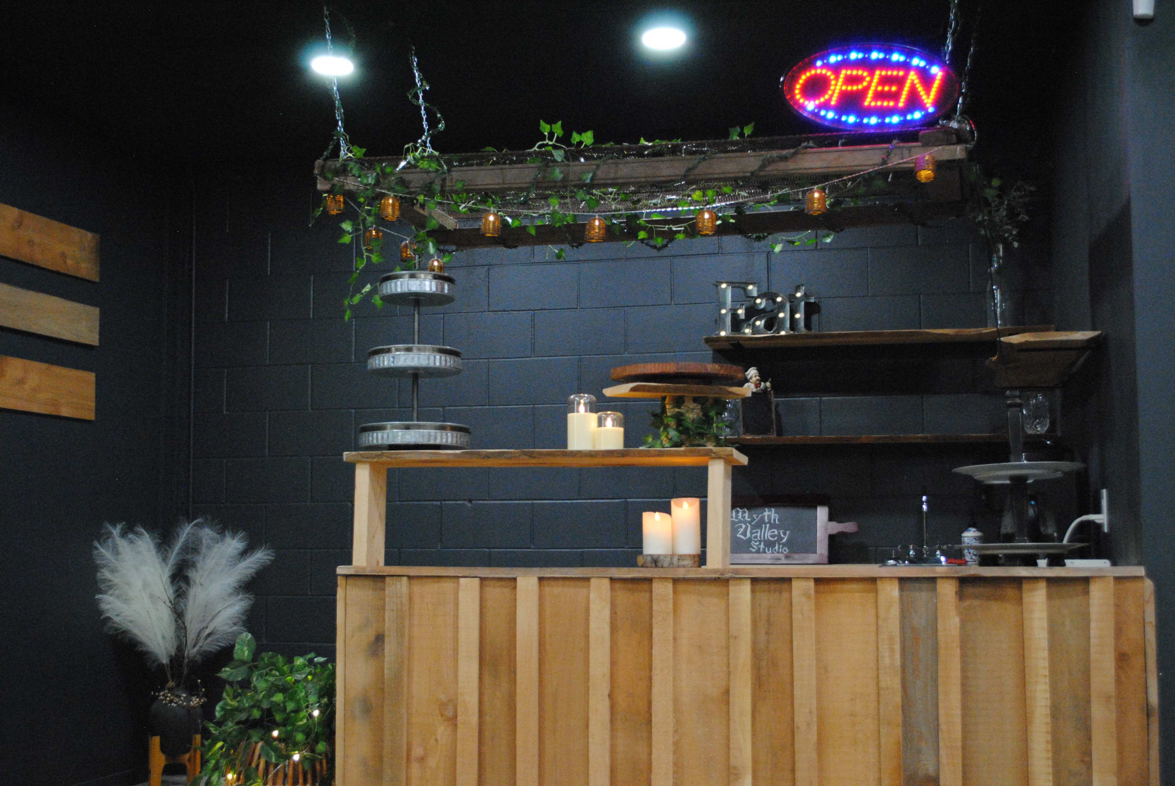 A wooden reception desk with an "OPEN" sign, surrounded by plants and decorative elements, is set against a dark wall in a welcoming space.