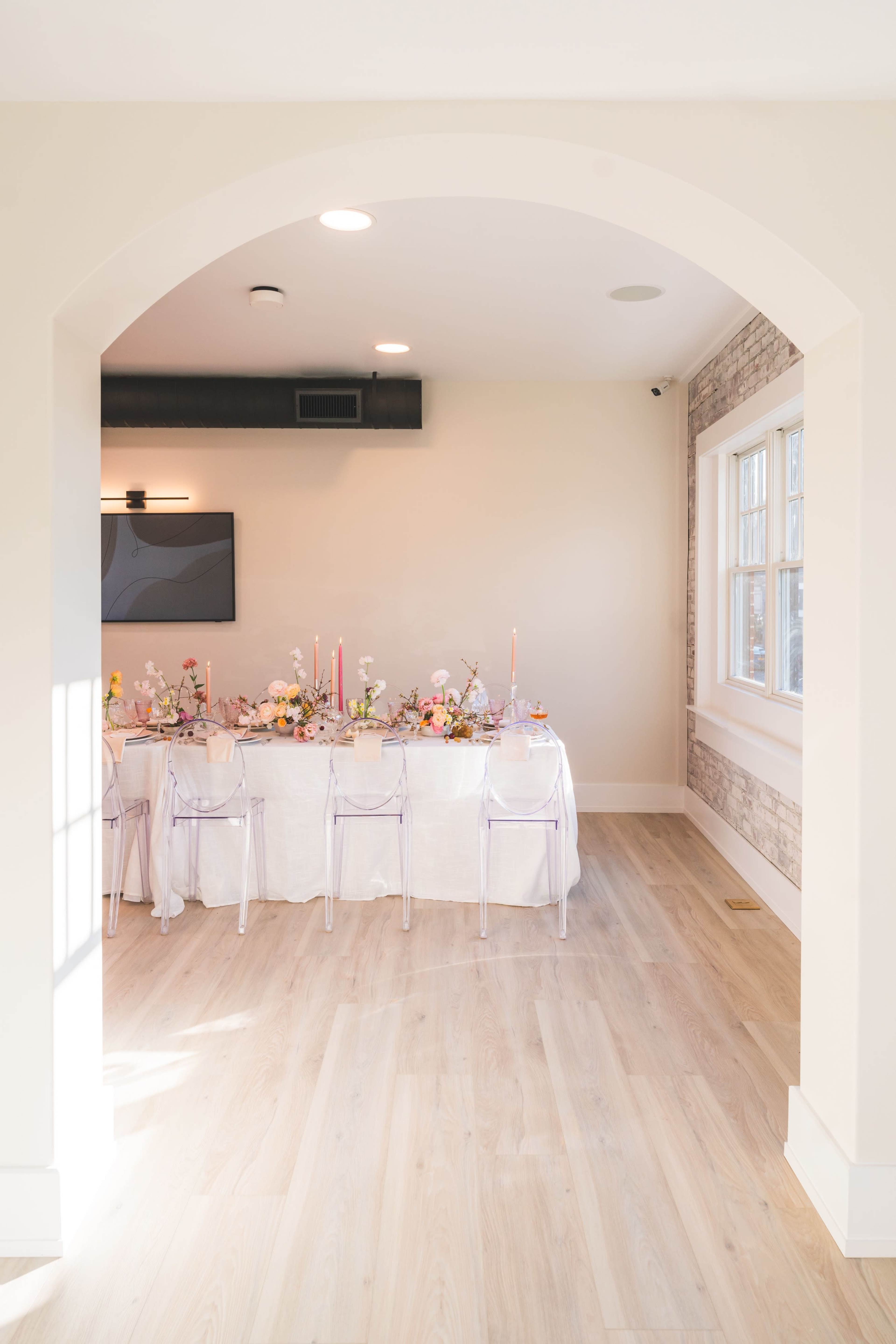 A dining area with a long table set for an event, featuring clear chairs and floral arrangements, is visible through an archway.