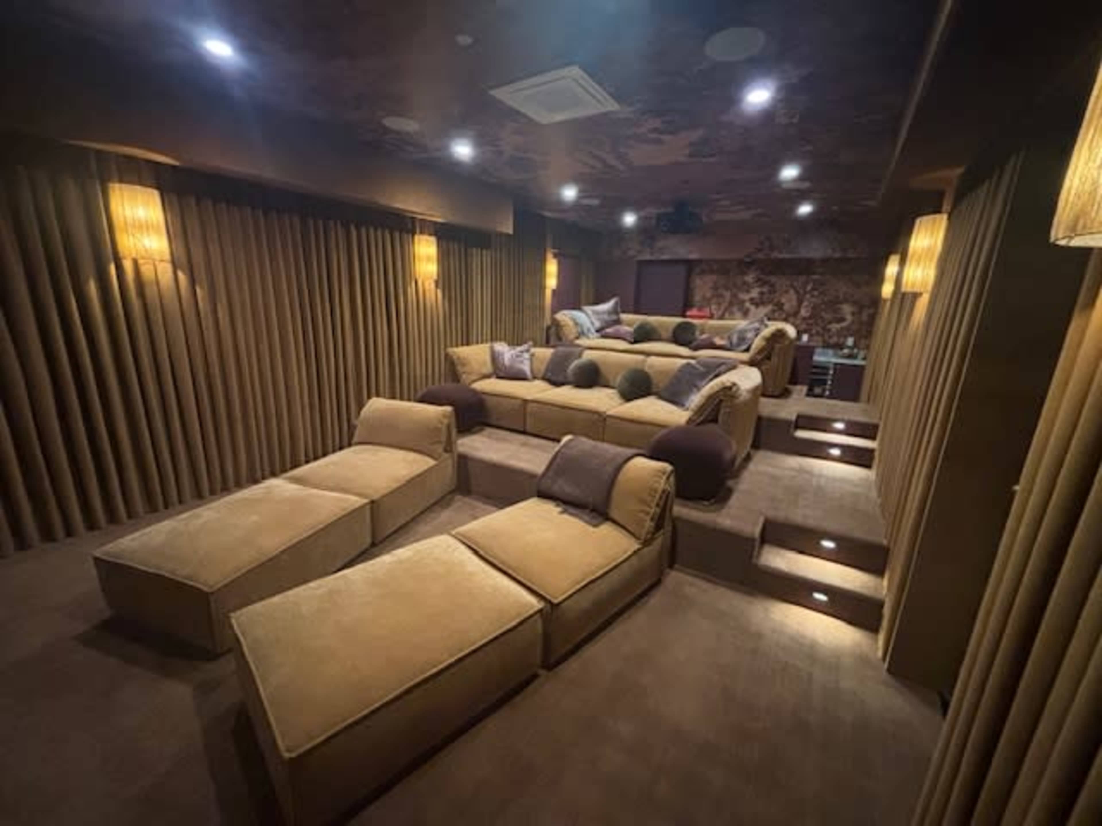 The image shows a home theater room with tiered seating, soft golden-toned fabric walls, and ambient lighting.