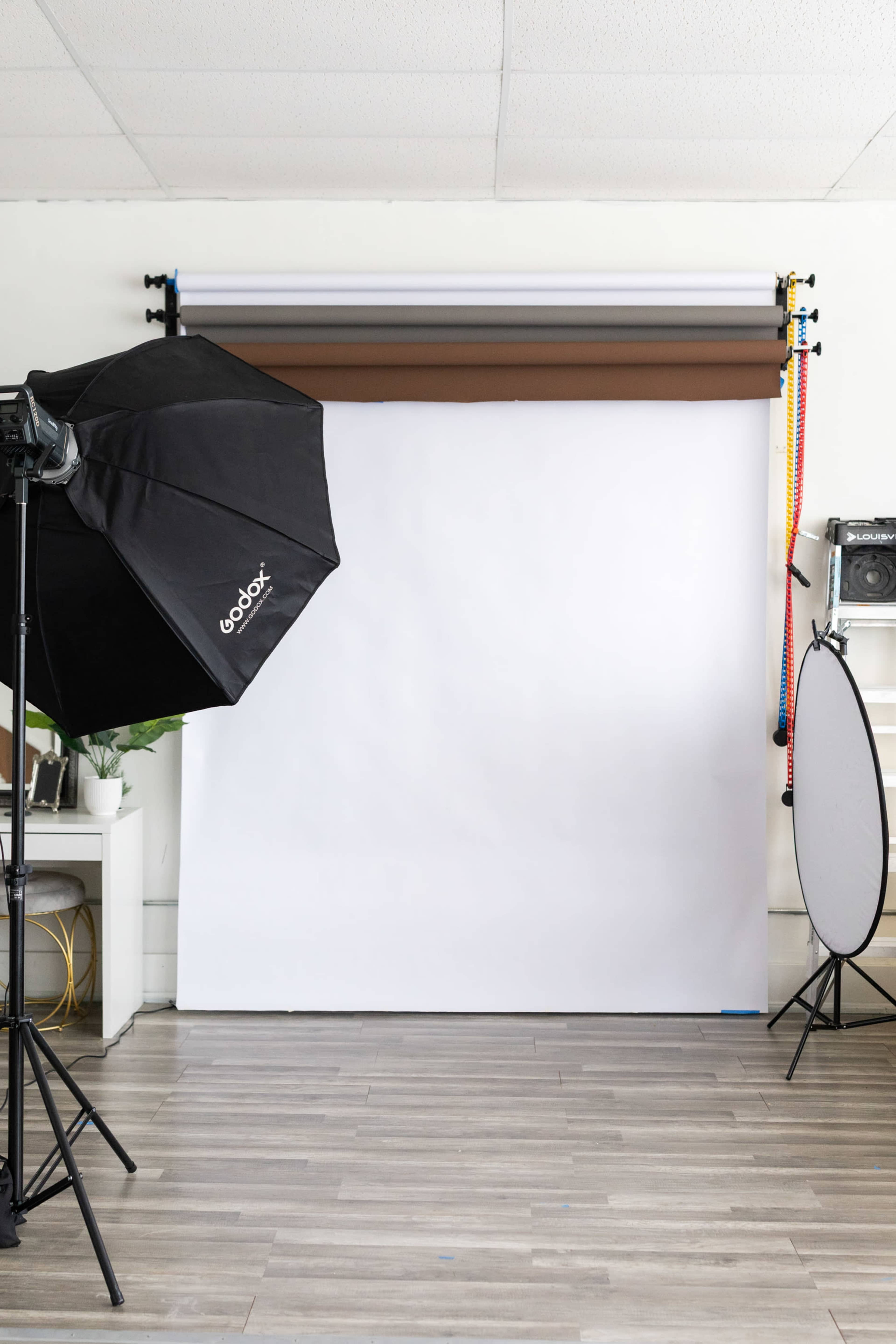 The image shows a photography studio setup featuring a white backdrop, softbox lighting, and a reflector.