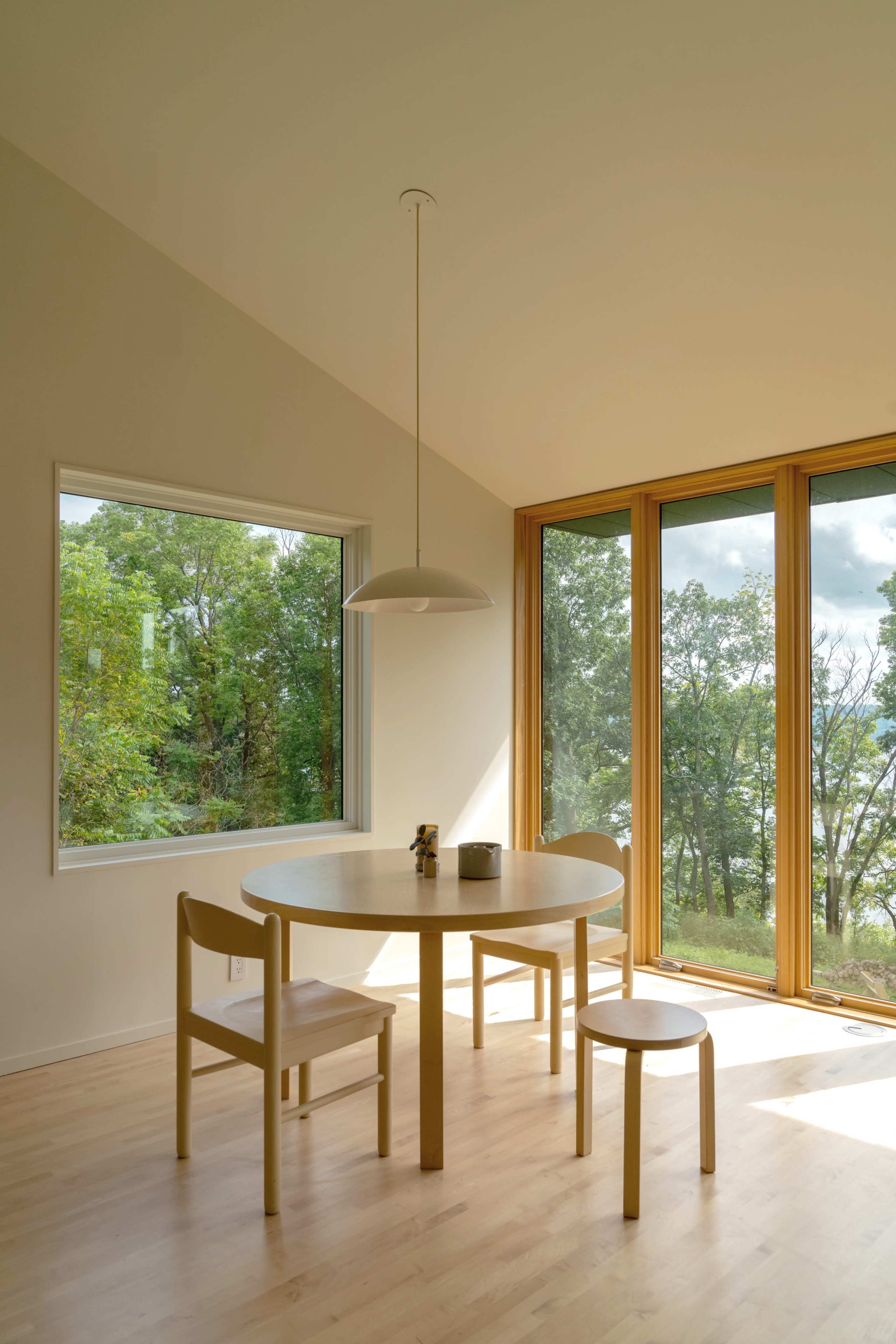 A small dining area with a round table and two chairs is situated beside large windows overlooking a forested area.