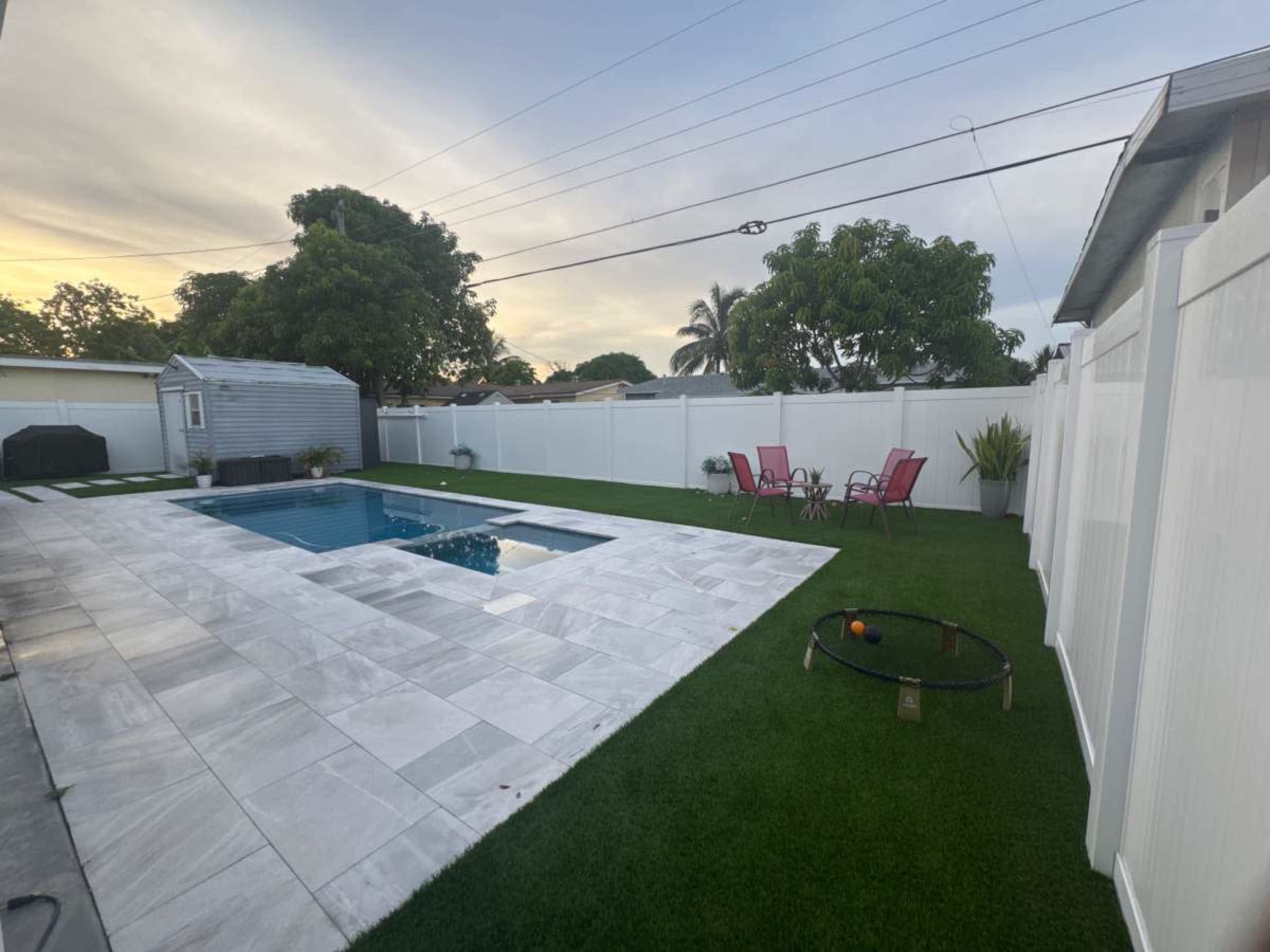 The image shows a backyard with a rectangular swimming pool, surrounded by marble tiles, artificial grass, and a seating area with red chairs.