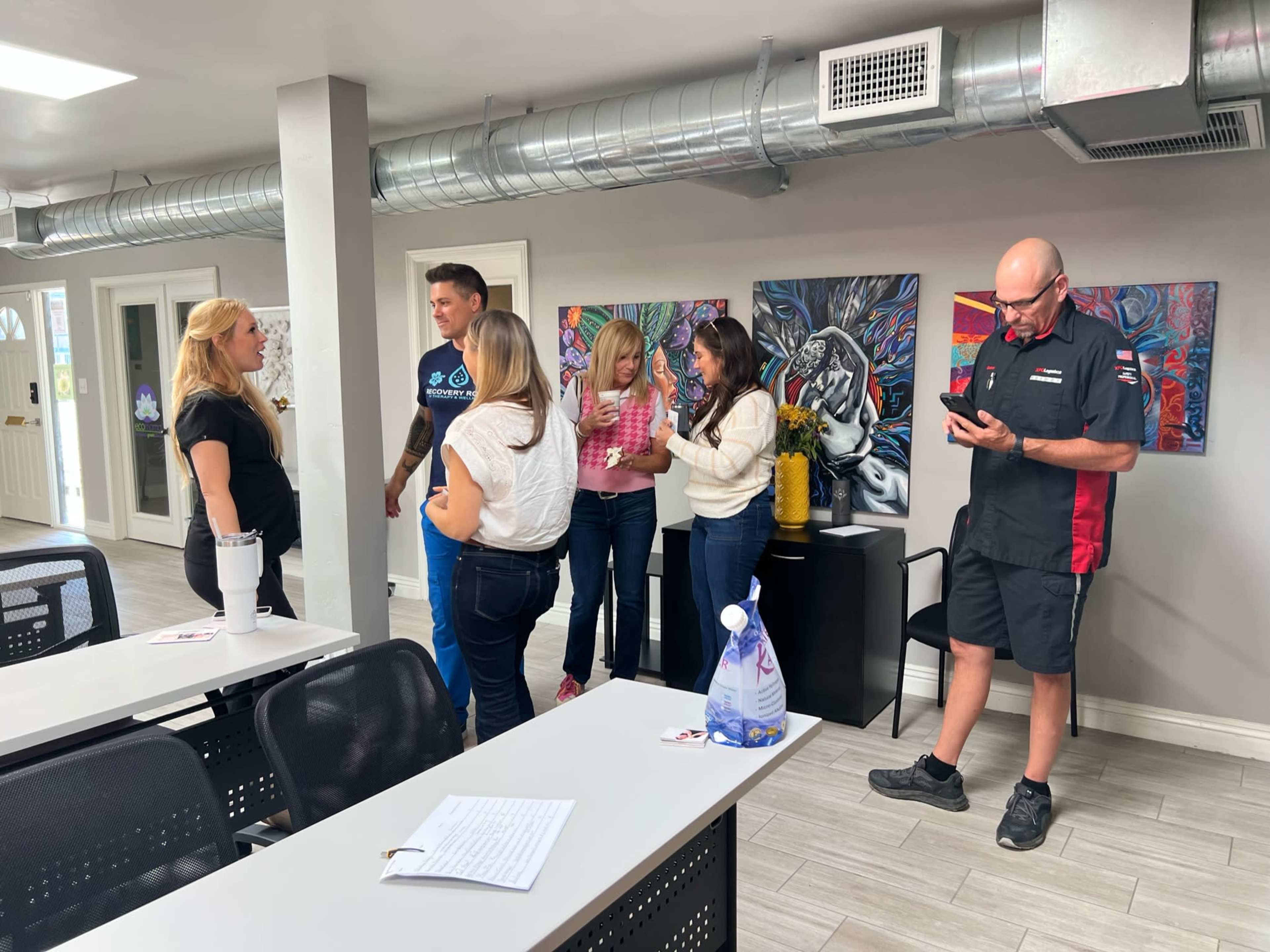A group of six people engaged in conversation stands in a modern office space with artwork on the walls and tables arranged for a meeting.