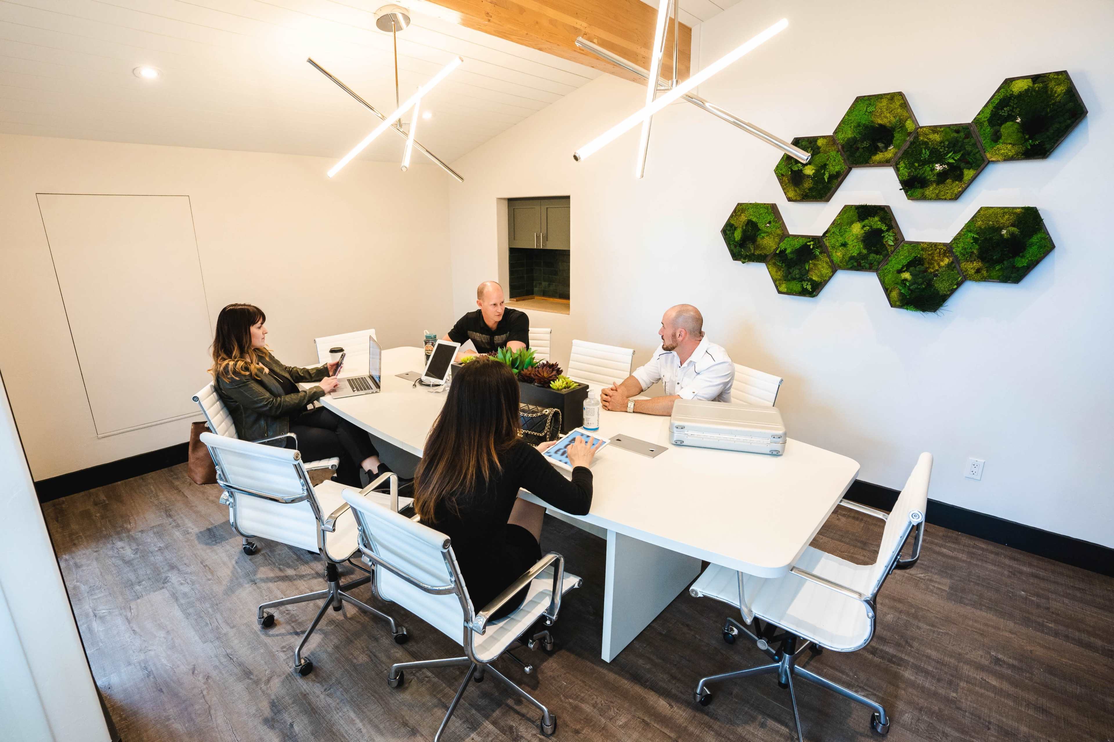 A sleek conference room features a large white table surrounded by four people engaged in discussion, with modern lighting and greenery on the wall.