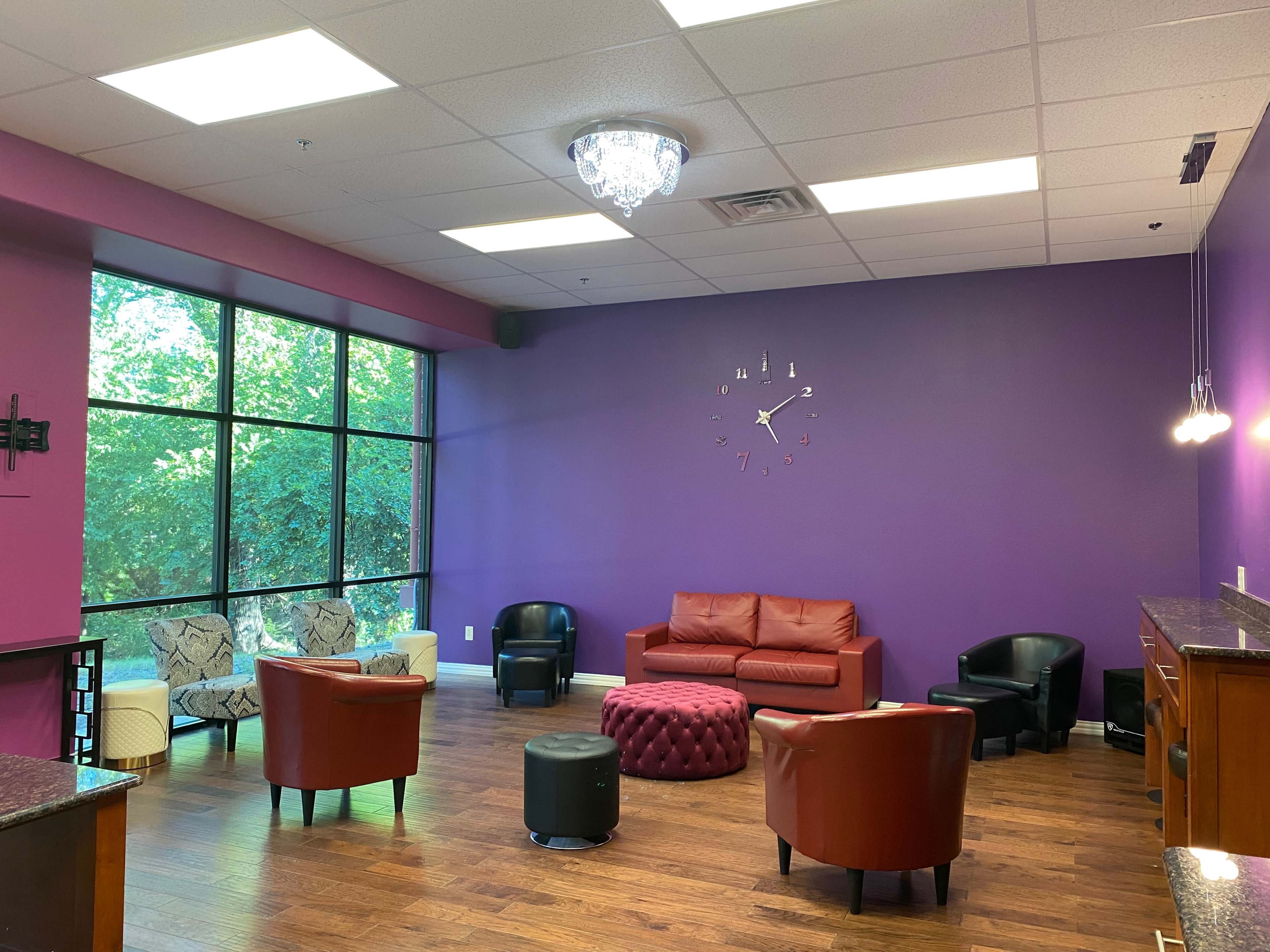 A modern lounge area with a purple wall, a mix of seating options including a red couch, armchairs, and a pouf, and large windows allowing natural light.
