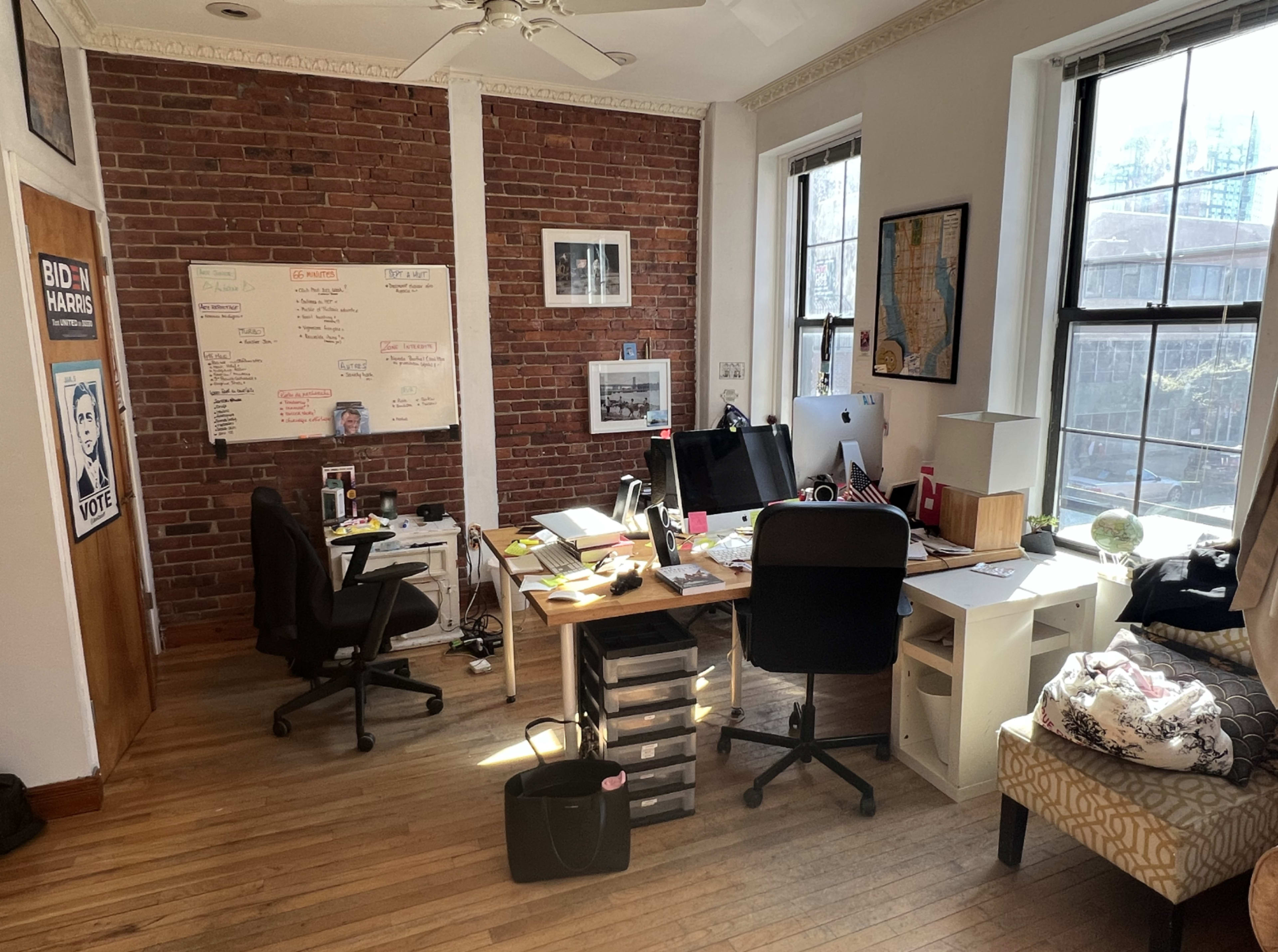 A modern office space with a wooden floor, brick walls, and a desk cluttered with various office supplies and electronic devices.