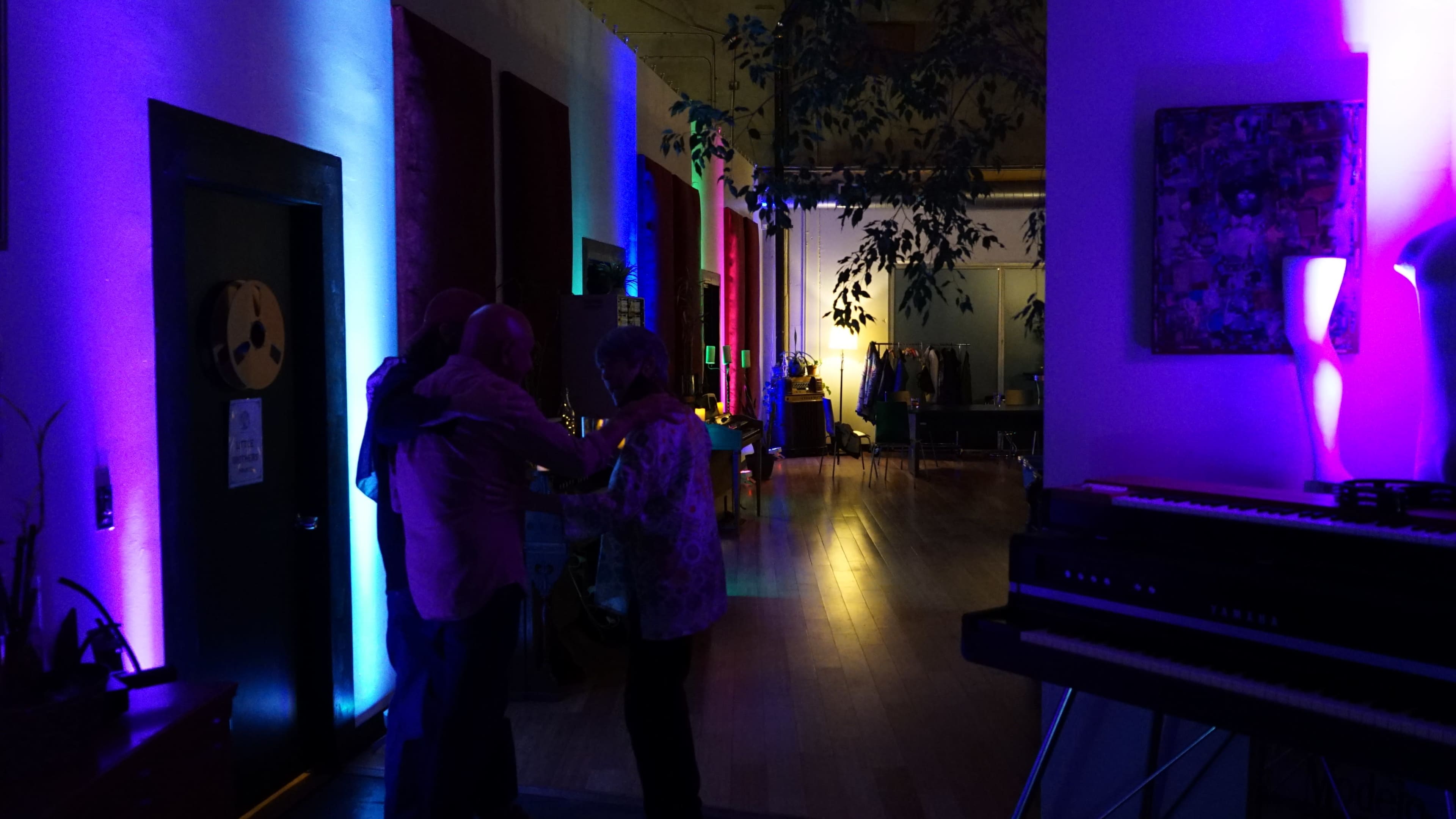 A brightly lit hallway with colorful lights features several people gathered near a piano on the right side.