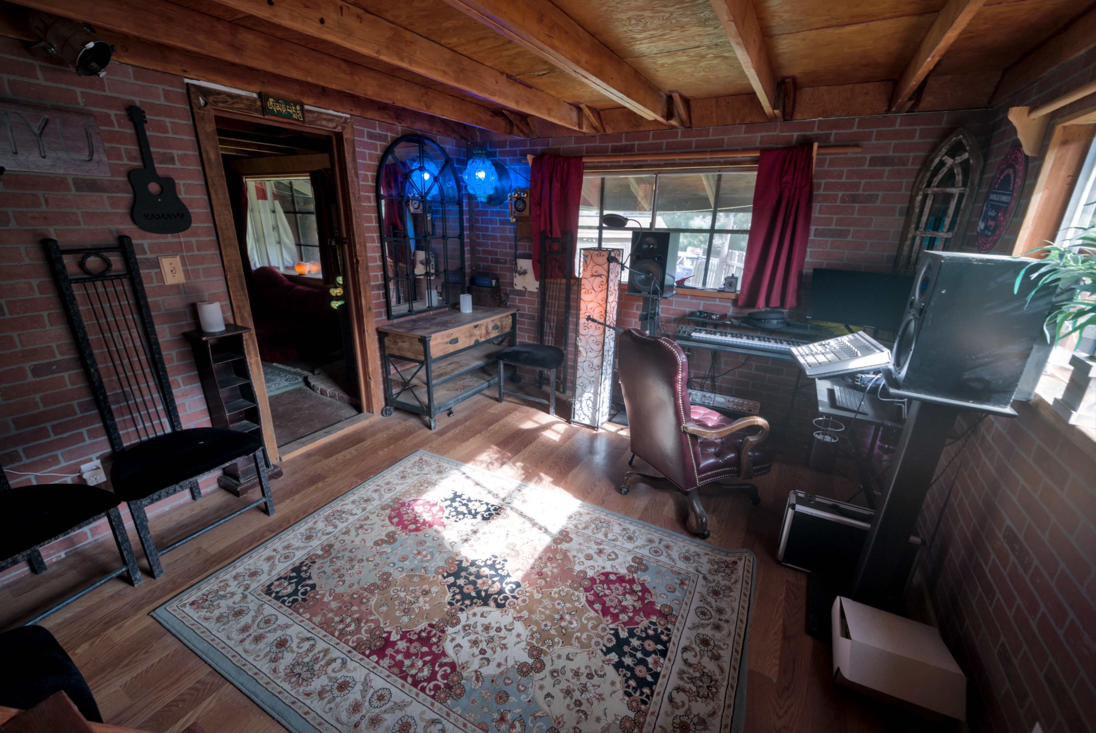 The image depicts a cozy studio workspace with brick walls, a wooden floor, and various musical instruments and equipment arranged around a vibrant area rug.