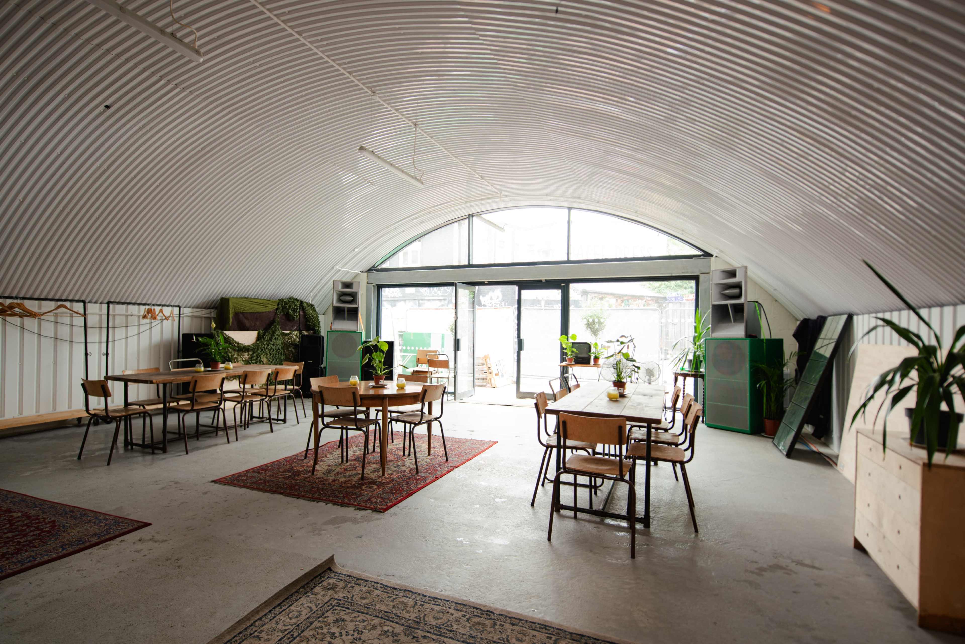 The image shows a spacious, curved interior with several wooden tables and chairs arranged in a light-filled room adorned with plants and rugs.