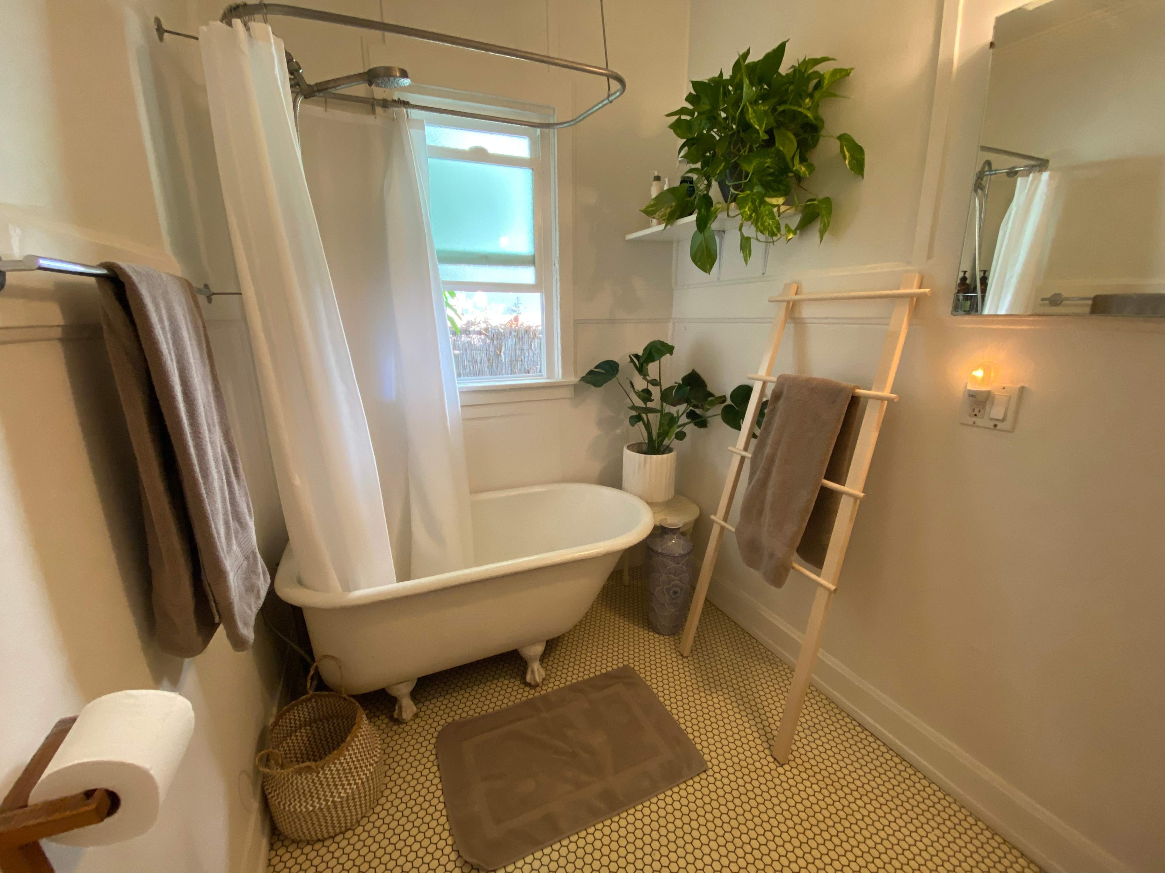 The image shows a small bathroom featuring a freestanding bathtub, a shower curtain, a sink with a mirror, and plants adding greenery to the space.