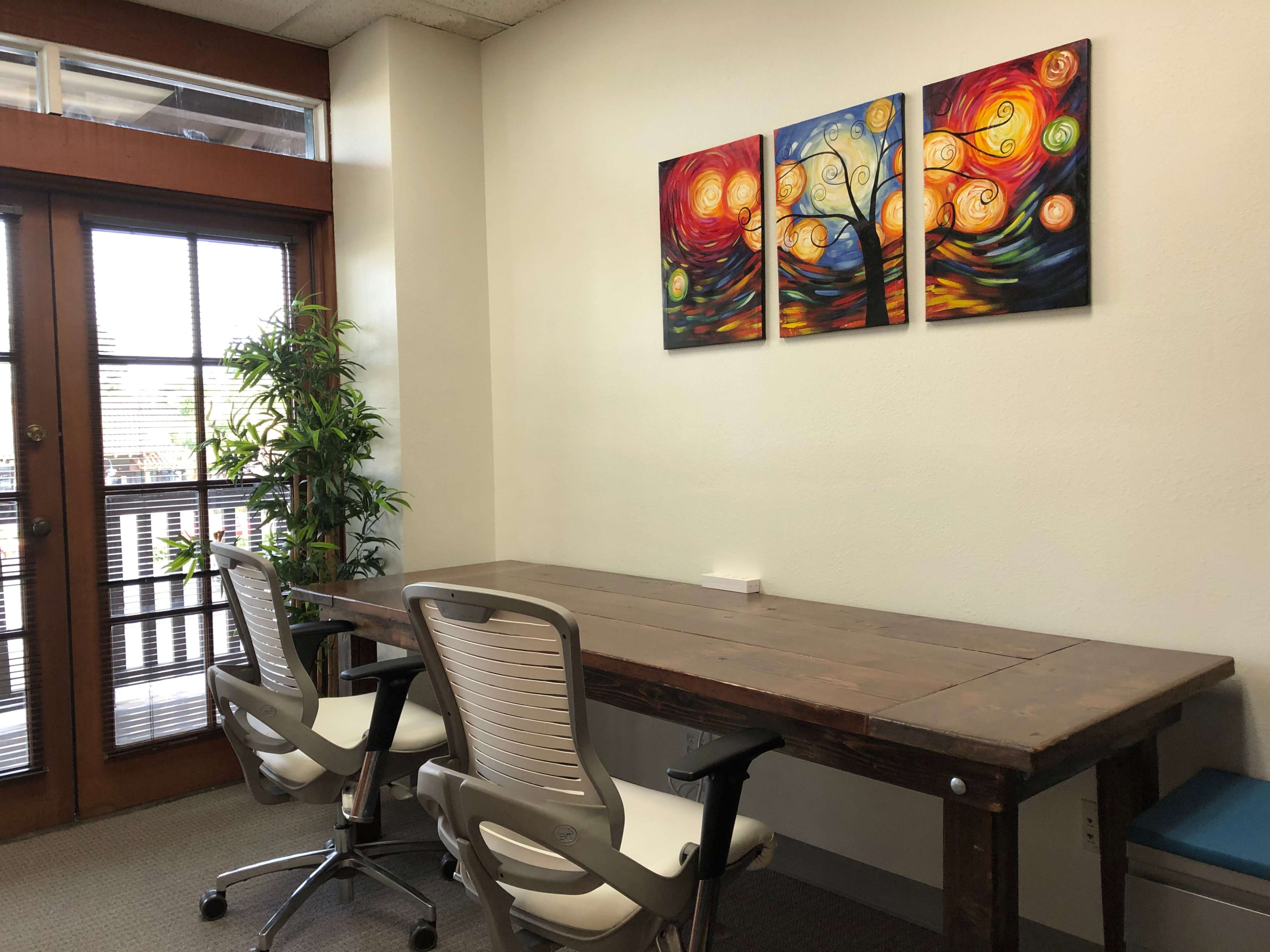 A minimalistic office space featuring a wooden table, two modern chairs, and vibrant abstract artwork on the wall.