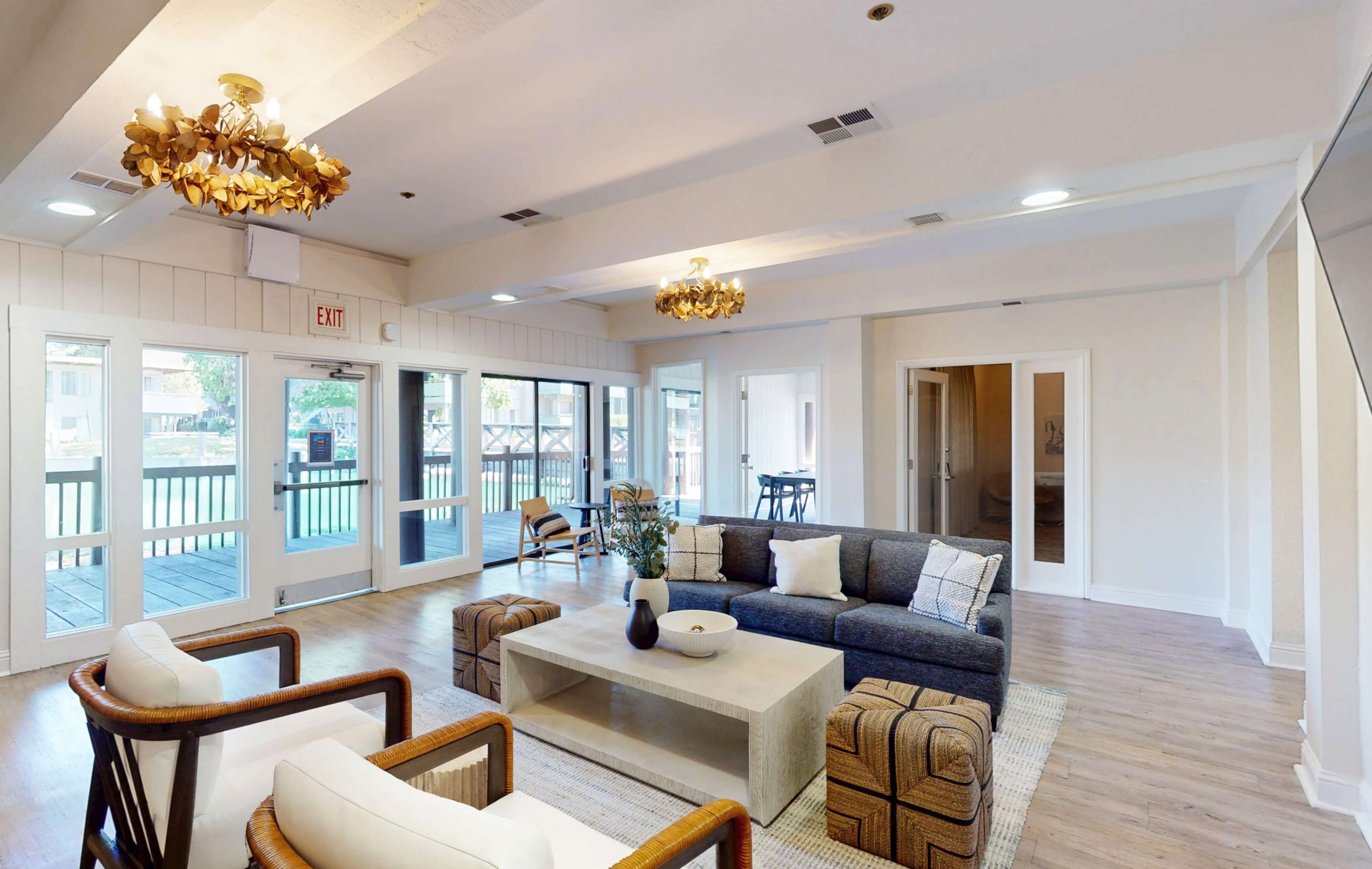 The image shows a well-lit lounge area featuring a gray sofa, two white armchairs, a coffee table, and large windows overlooking a deck.