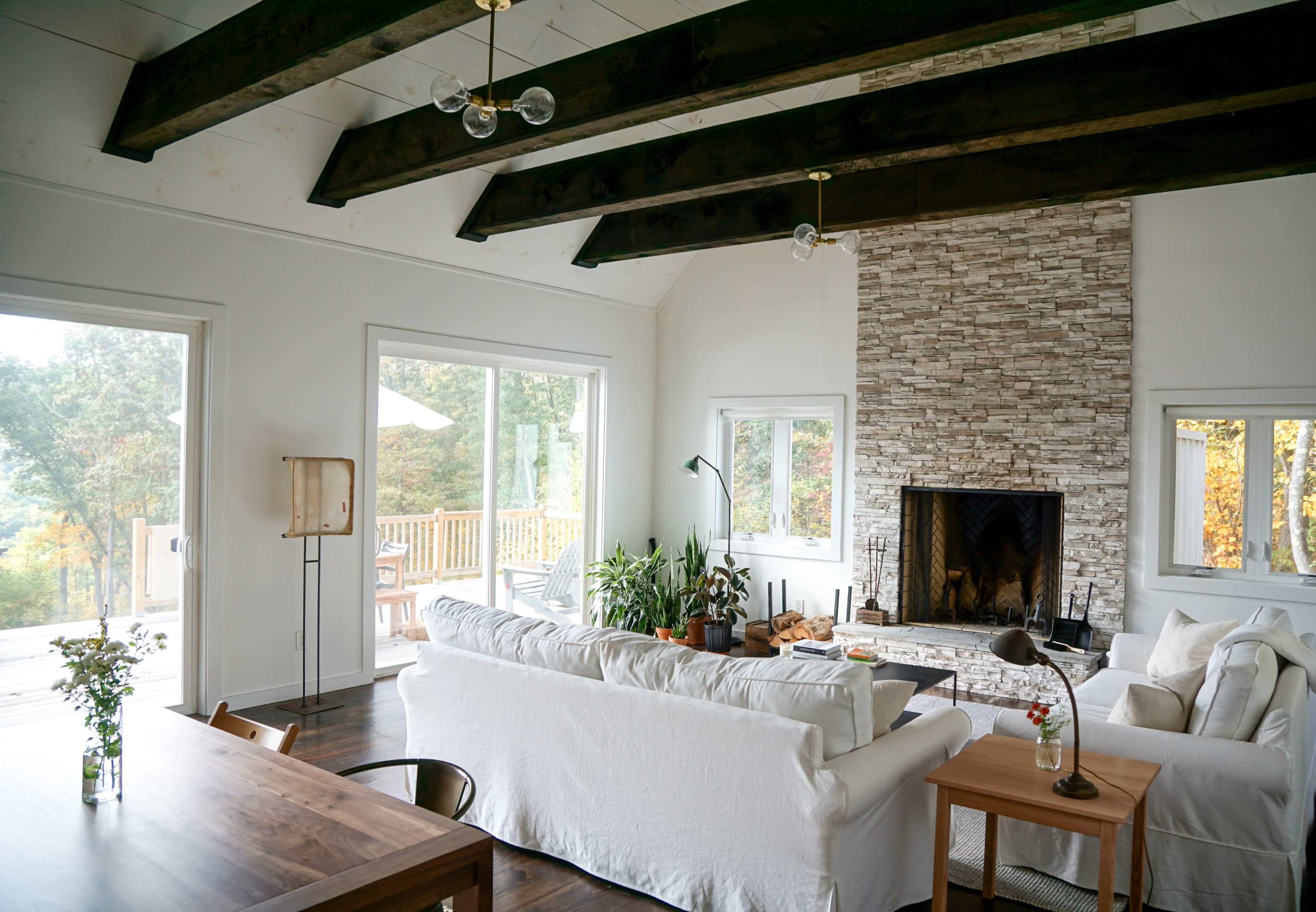 A modern living room with a stone fireplace, large windows, and wooden beams across the ceiling.