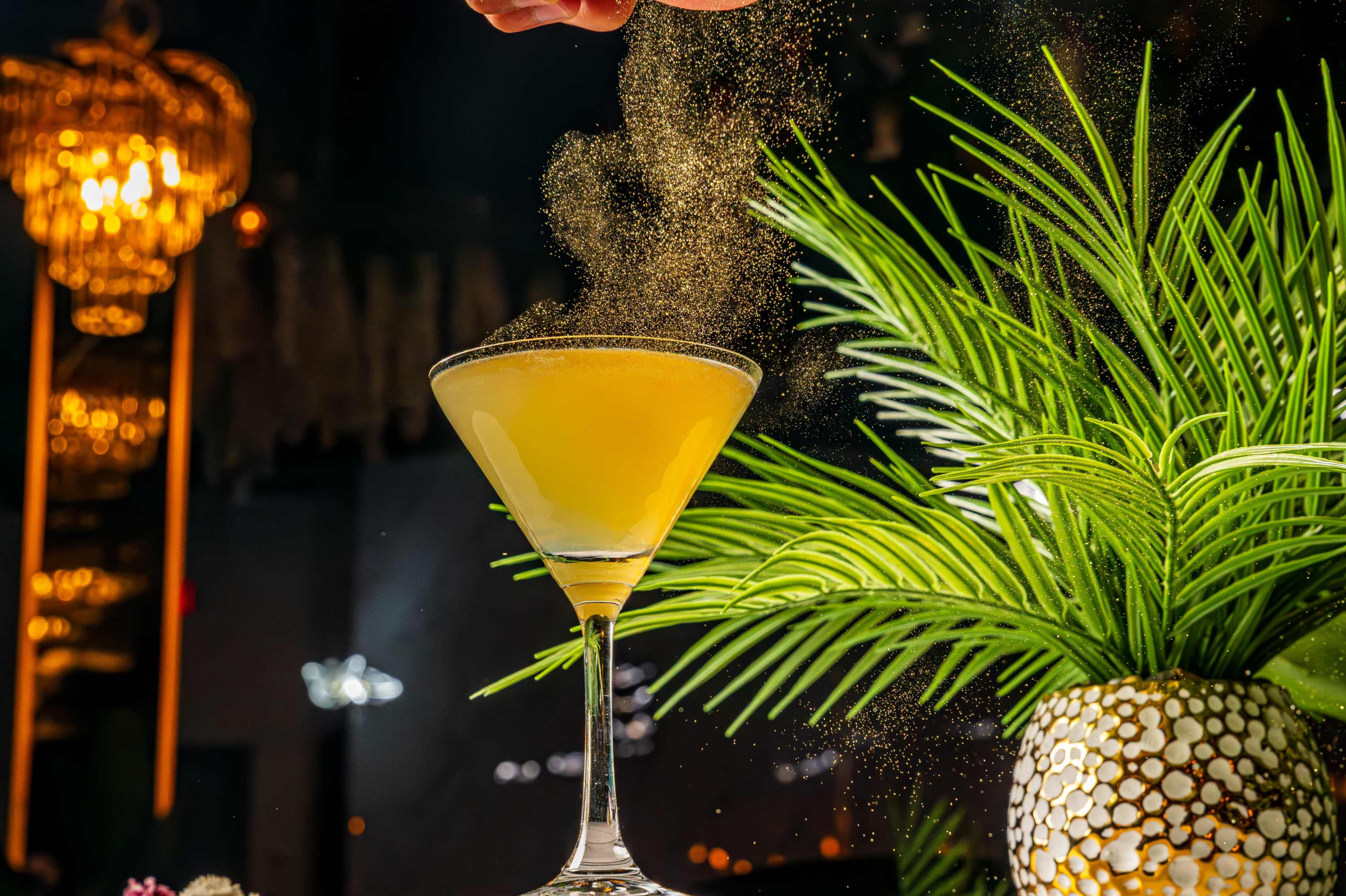 A hand sprinkles fine powder over a yellow cocktail in a martini glass, with greenery and decorative lighting in the background.