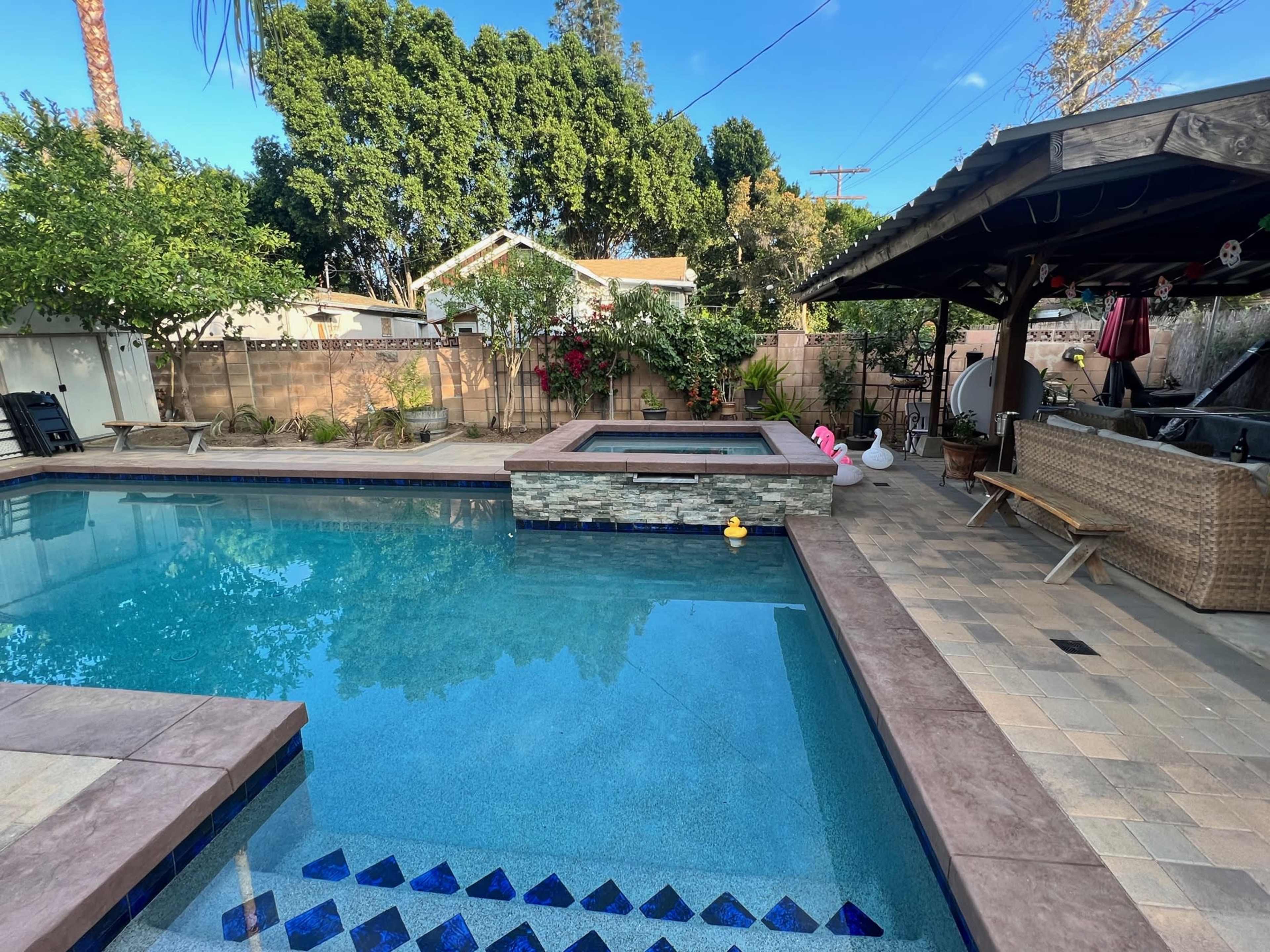 A backyard features a swimming pool with a spa, surrounded by stone coping, patio furniture, and landscaped greenery.