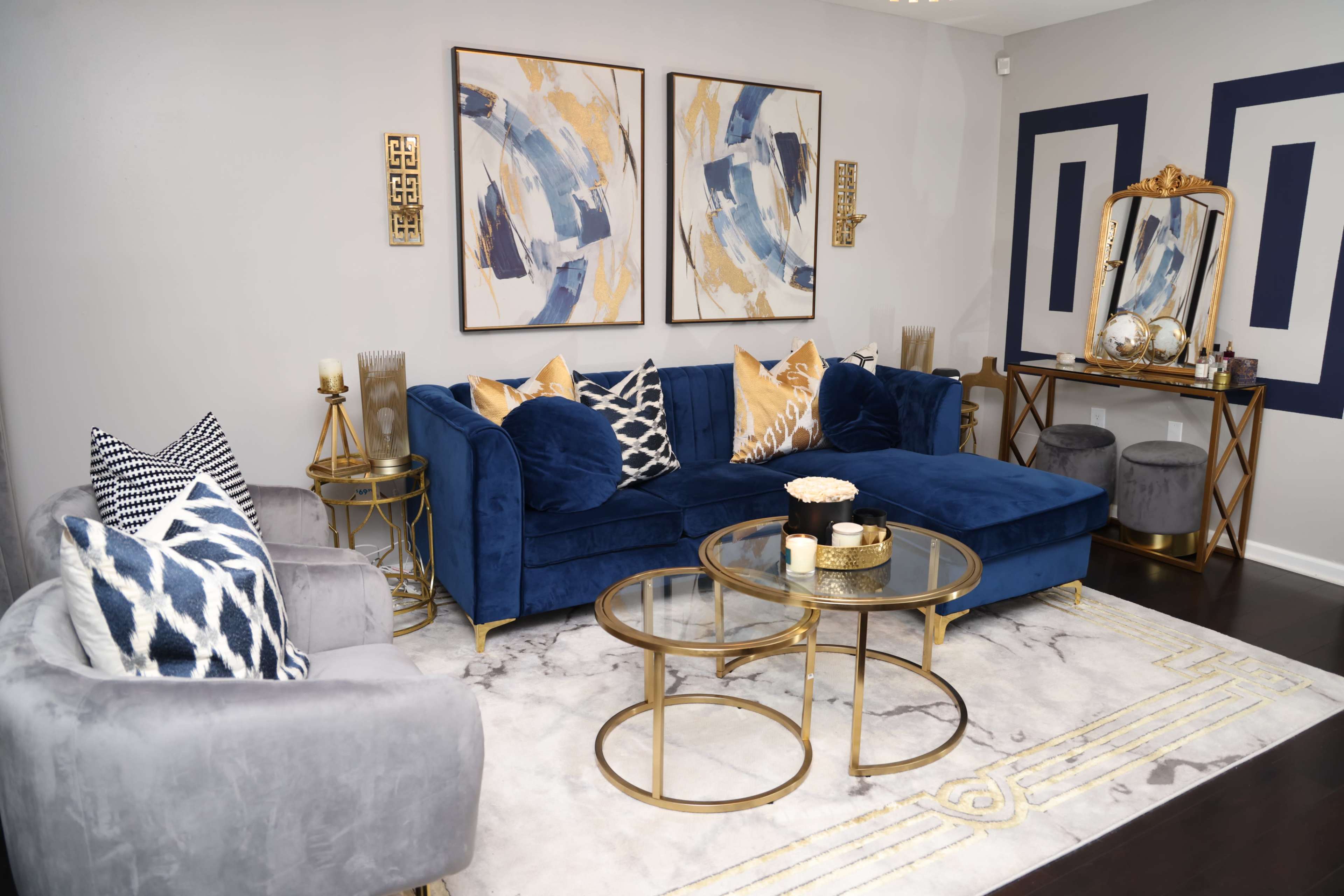 The image shows a modern living room featuring a blue velvet sofa, two decorative chairs, a coffee table, and abstract wall art, all arranged on a patterned rug.