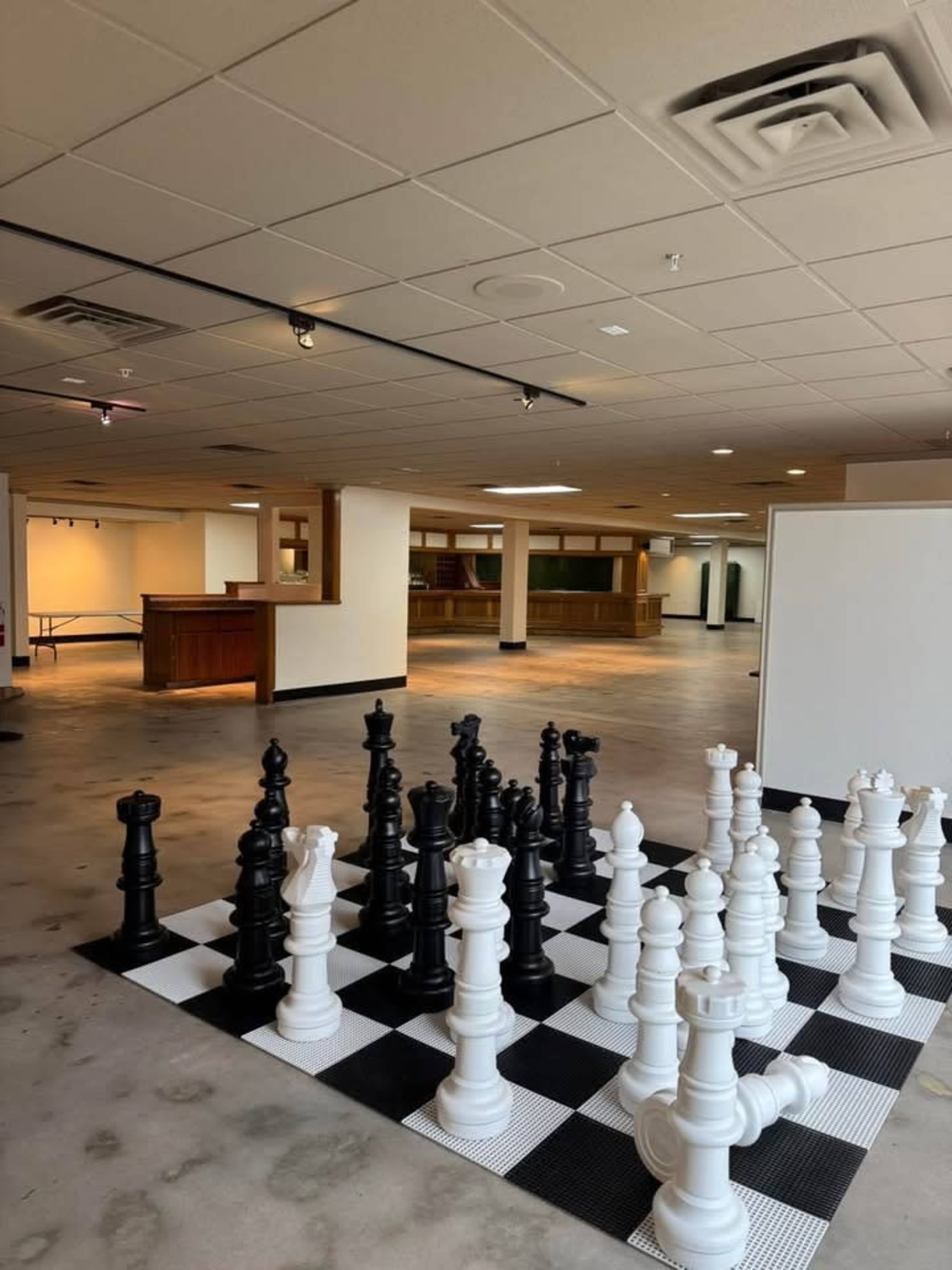 The image shows a large indoor space featuring a giant chess set with black and white pieces on a checkered floor.