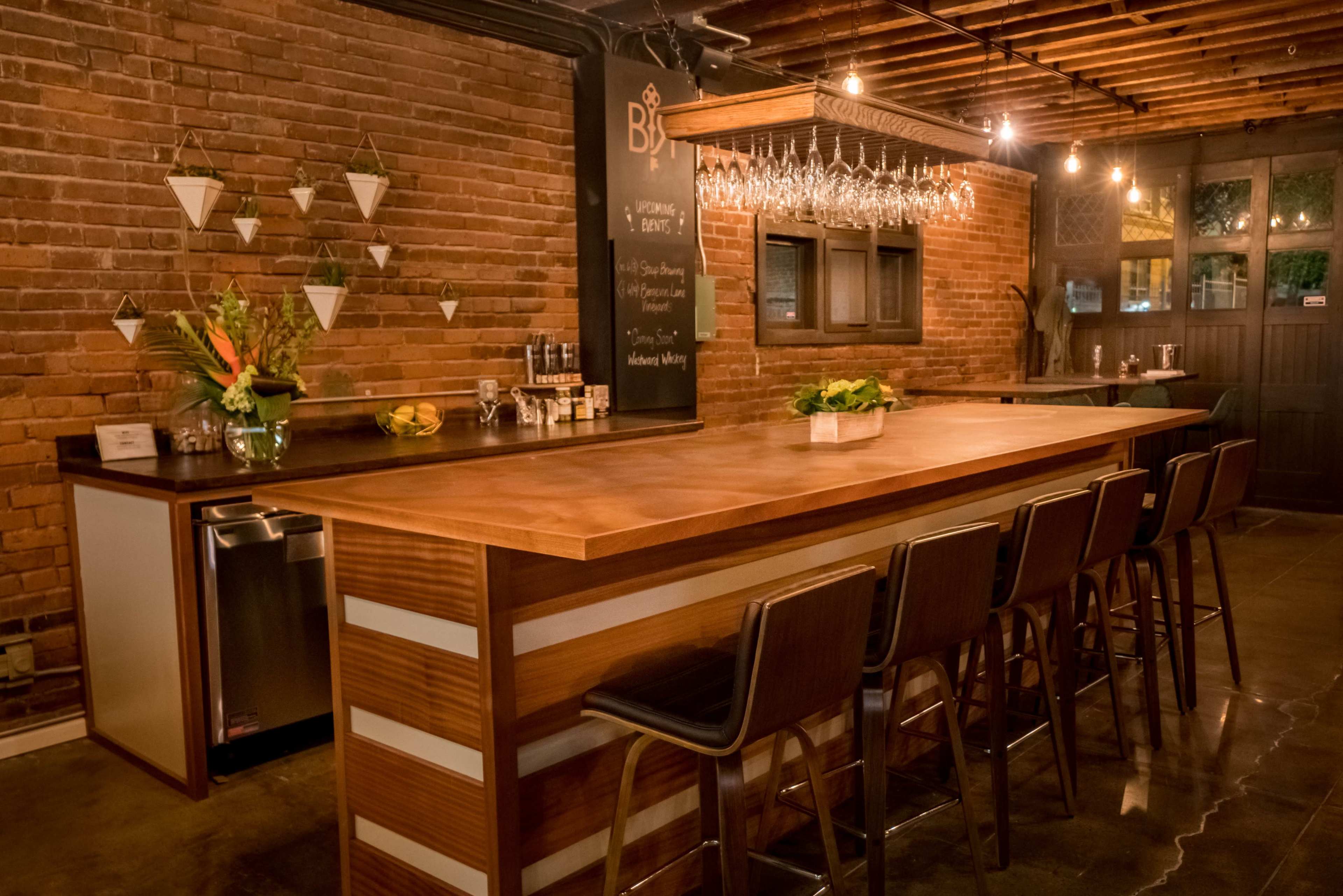 The image shows a modern bar area with a wooden countertop, high-backed chairs, and exposed brick walls, illuminated by hanging lights.