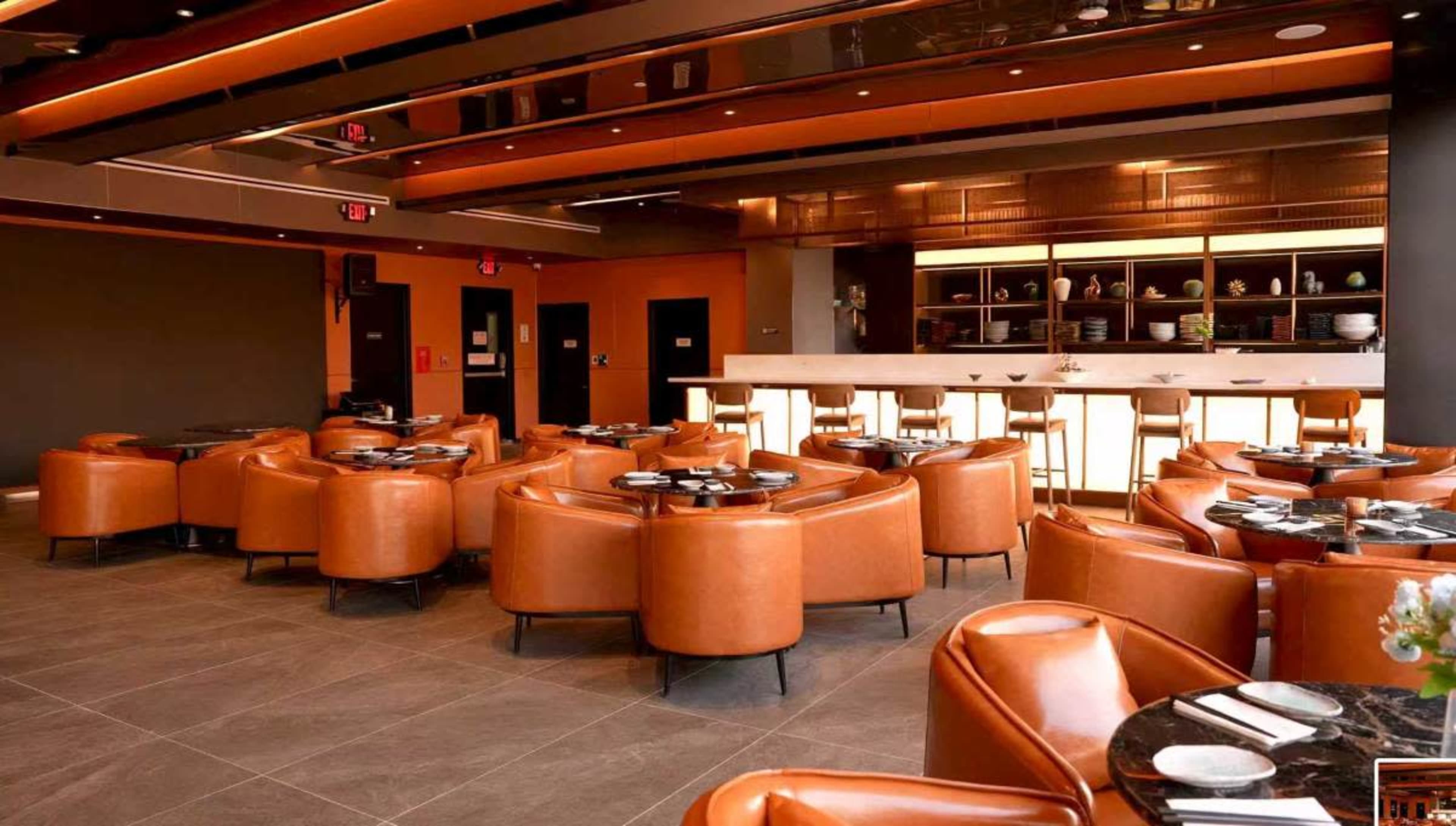 The image shows a modern restaurant interior featuring brown leather seating, a bar with high stools, and a well-lit open area.