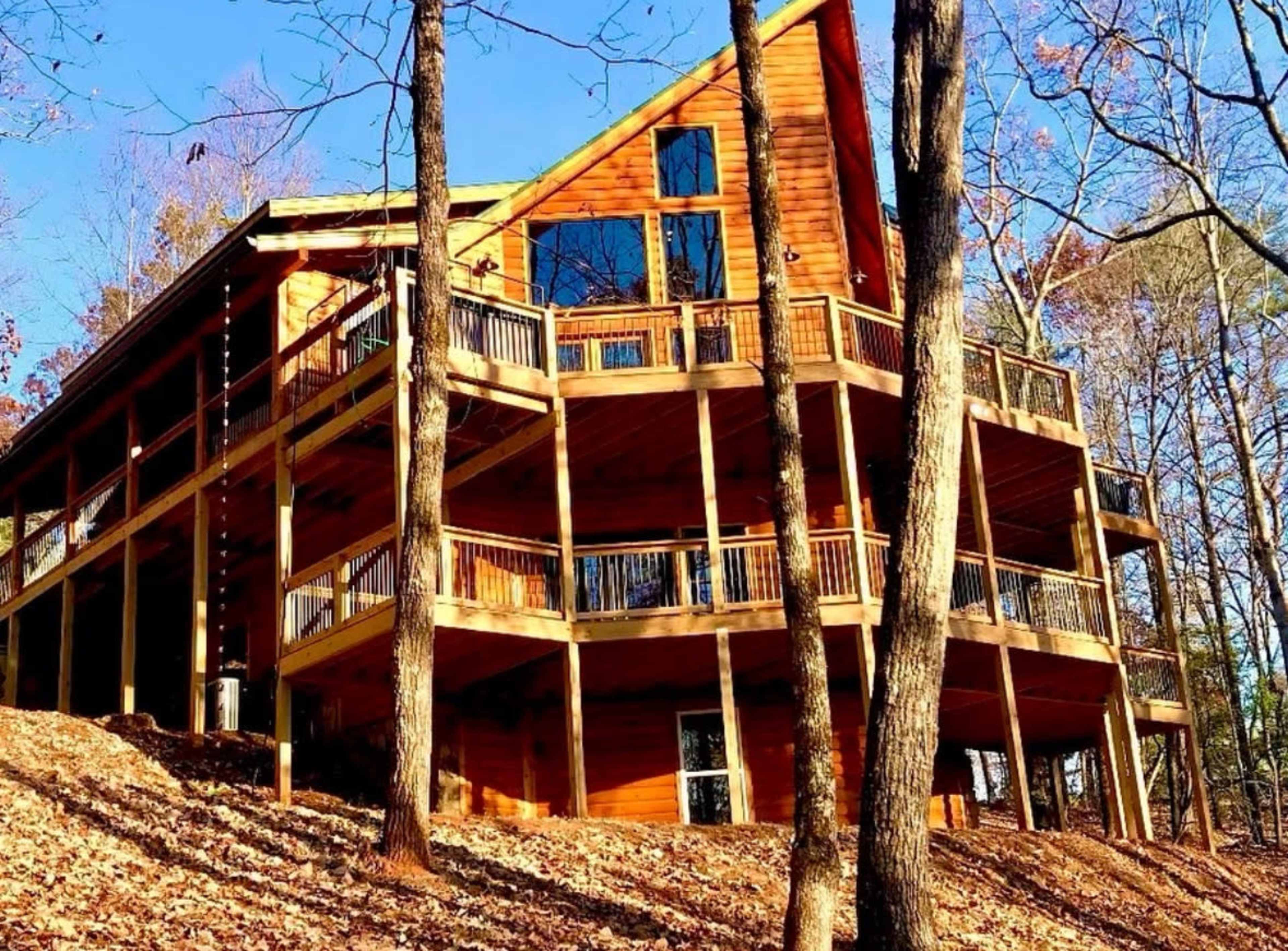 The image shows a large wooden cabin with multiple decks, situated on a hillside surrounded by trees and bare branches.