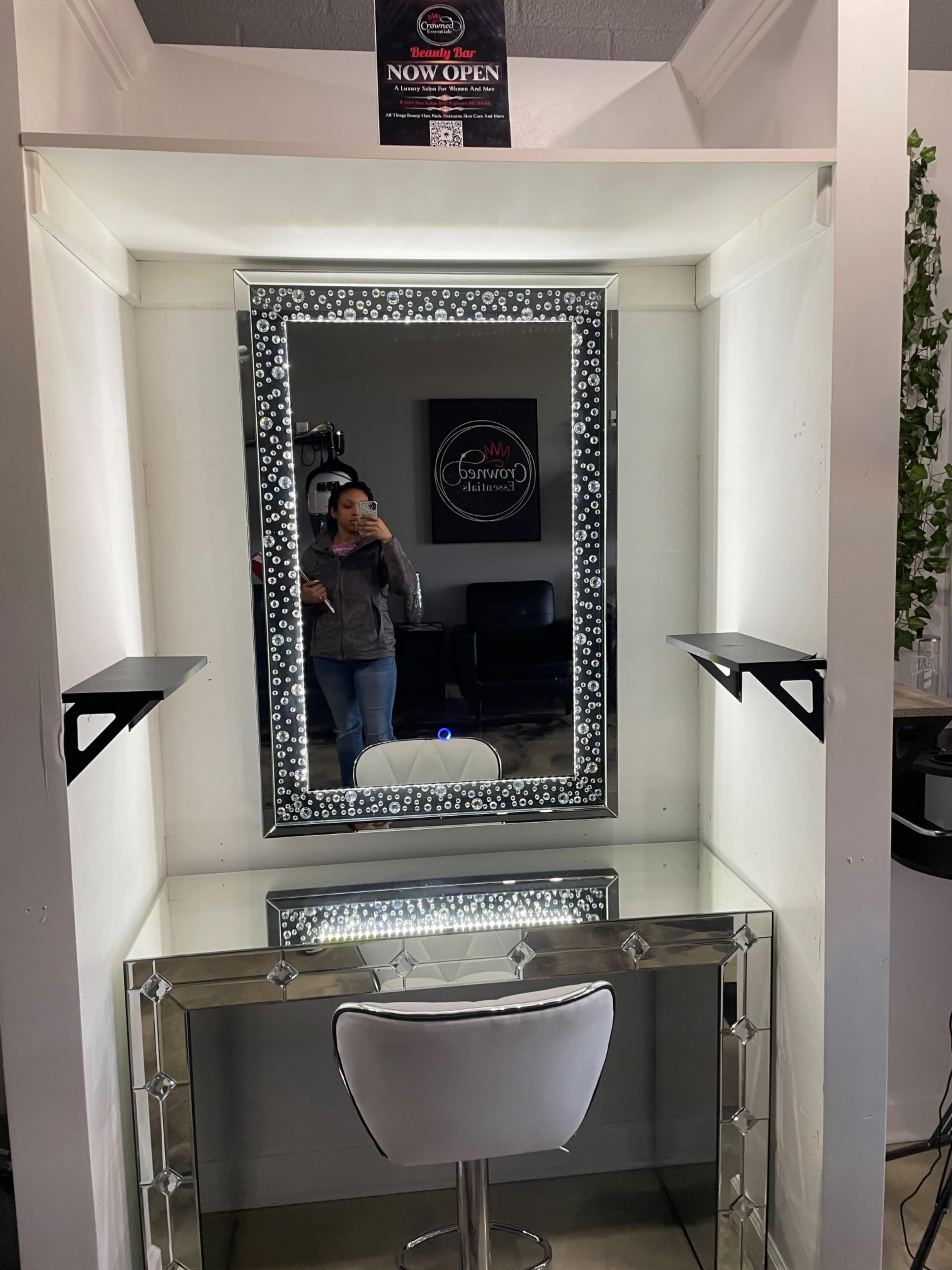 A salon vanity with a large mirrored frame, a white chair, and wall-mounted shelves on either side of the mirror.