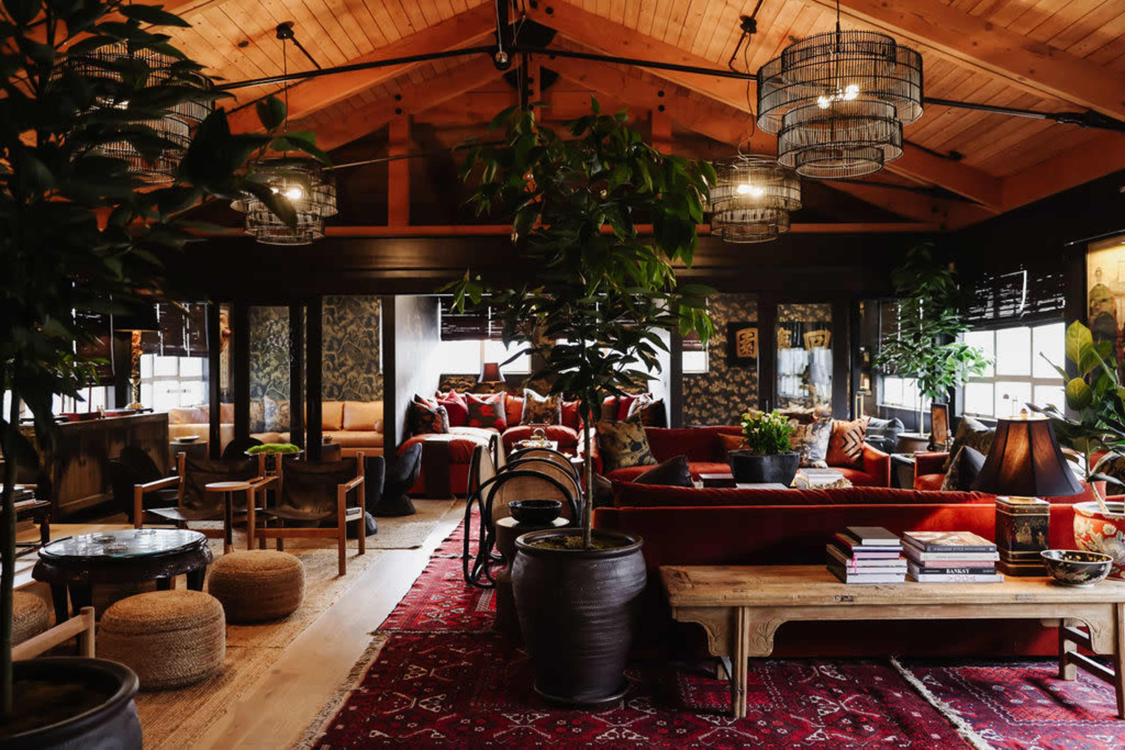 A spacious, wood-beamed living room decorated with plants, a large red sofa, and various tables under unique light fixtures.