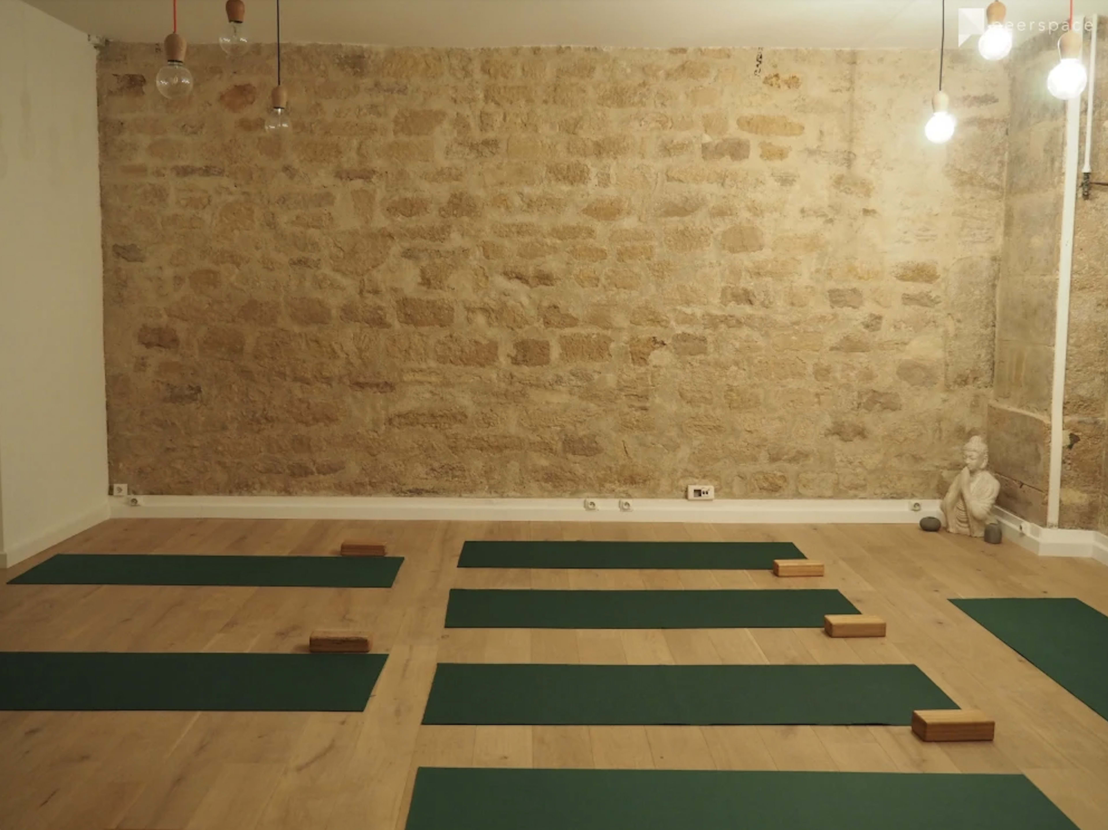 A spacious yoga studio with wooden flooring, a stone wall, and several green yoga mats arranged on the floor alongside wooden blocks.