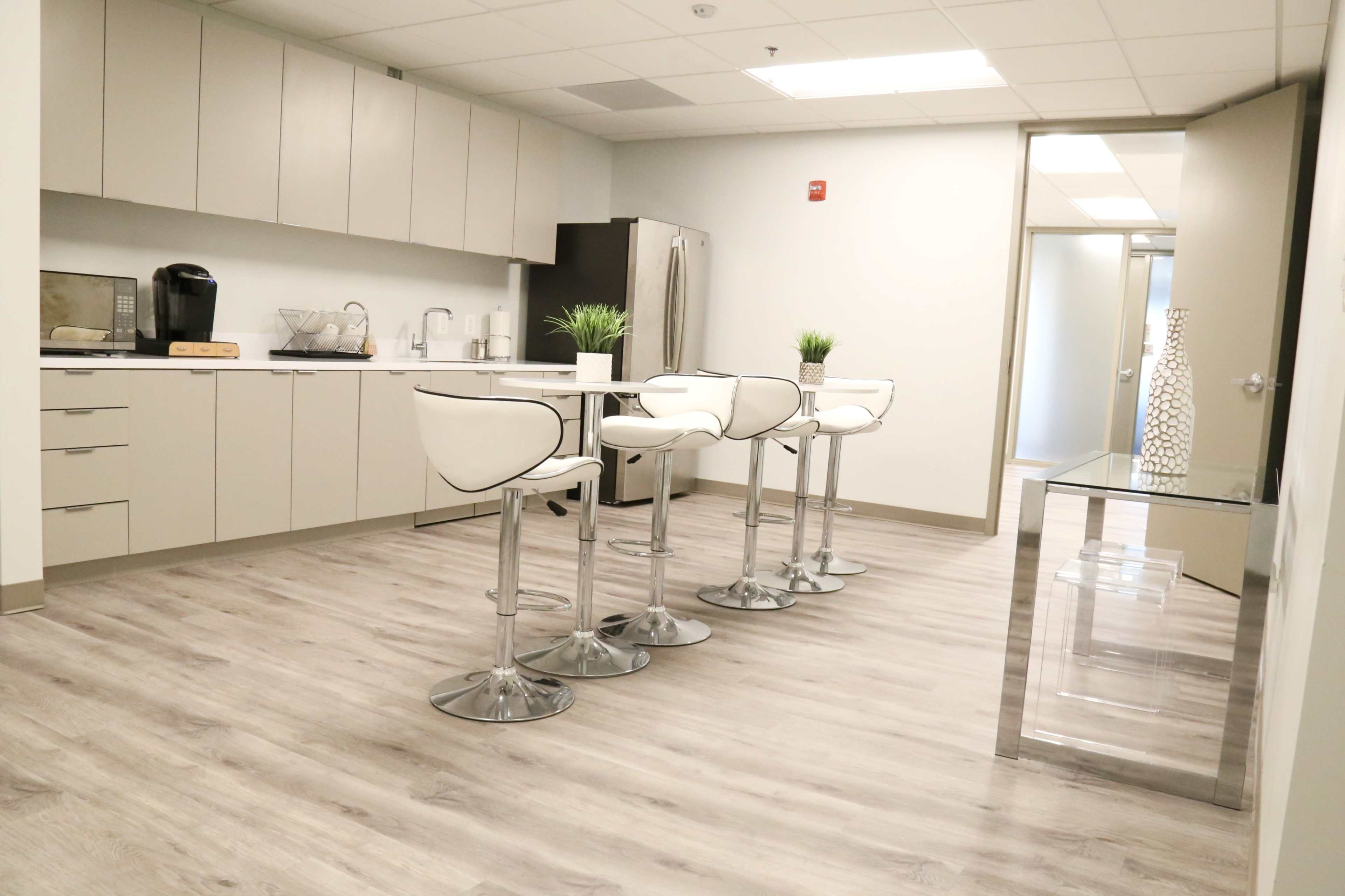 A modern kitchen area featuring a table with four white barstools, a coffee maker, and a stainless steel refrigerator against a light-colored cabinetry backdrop.