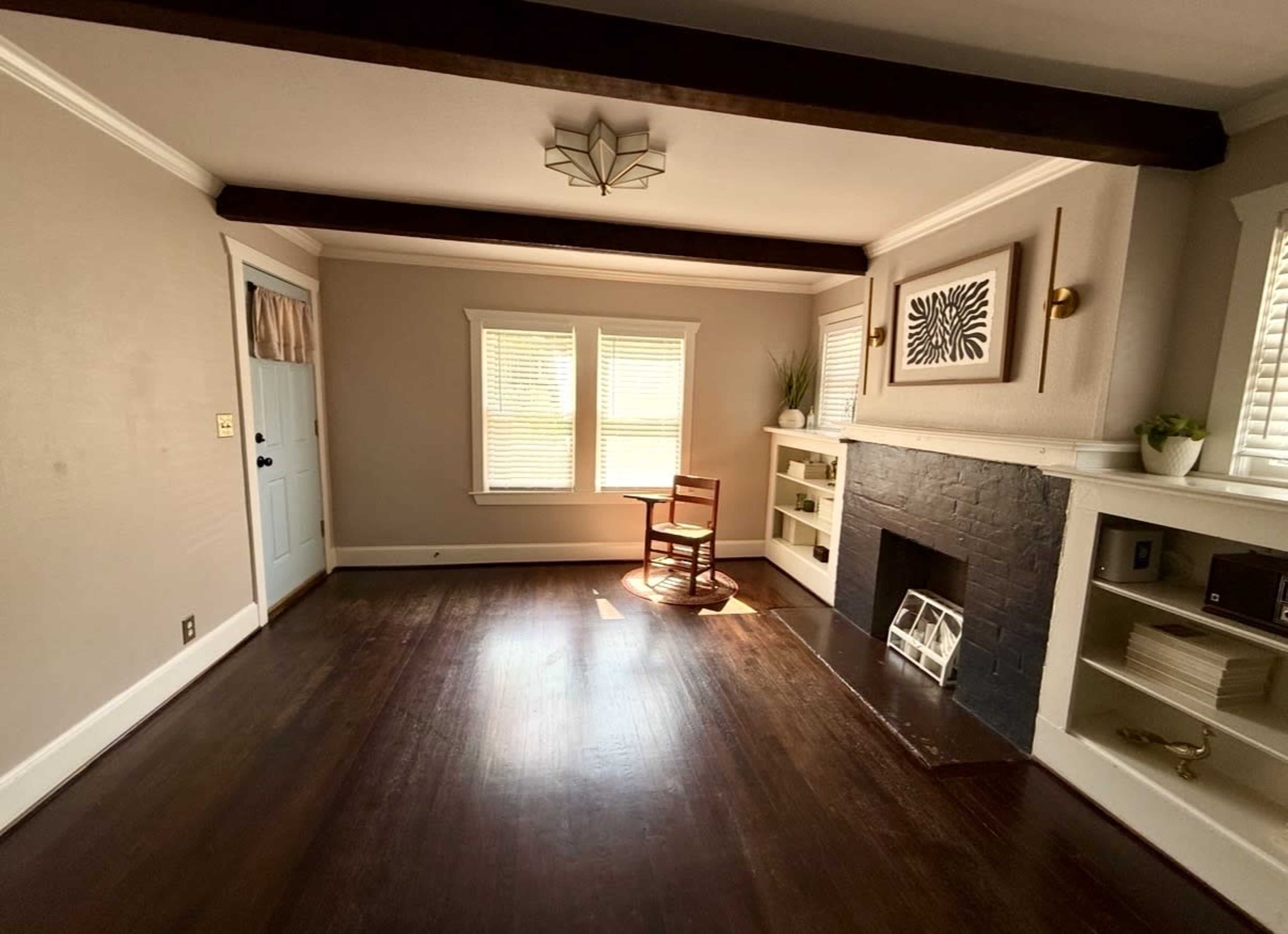The image shows a sparsely furnished living room featuring a wooden chair, a small table near the window, and a decorative fireplace with shelves.
