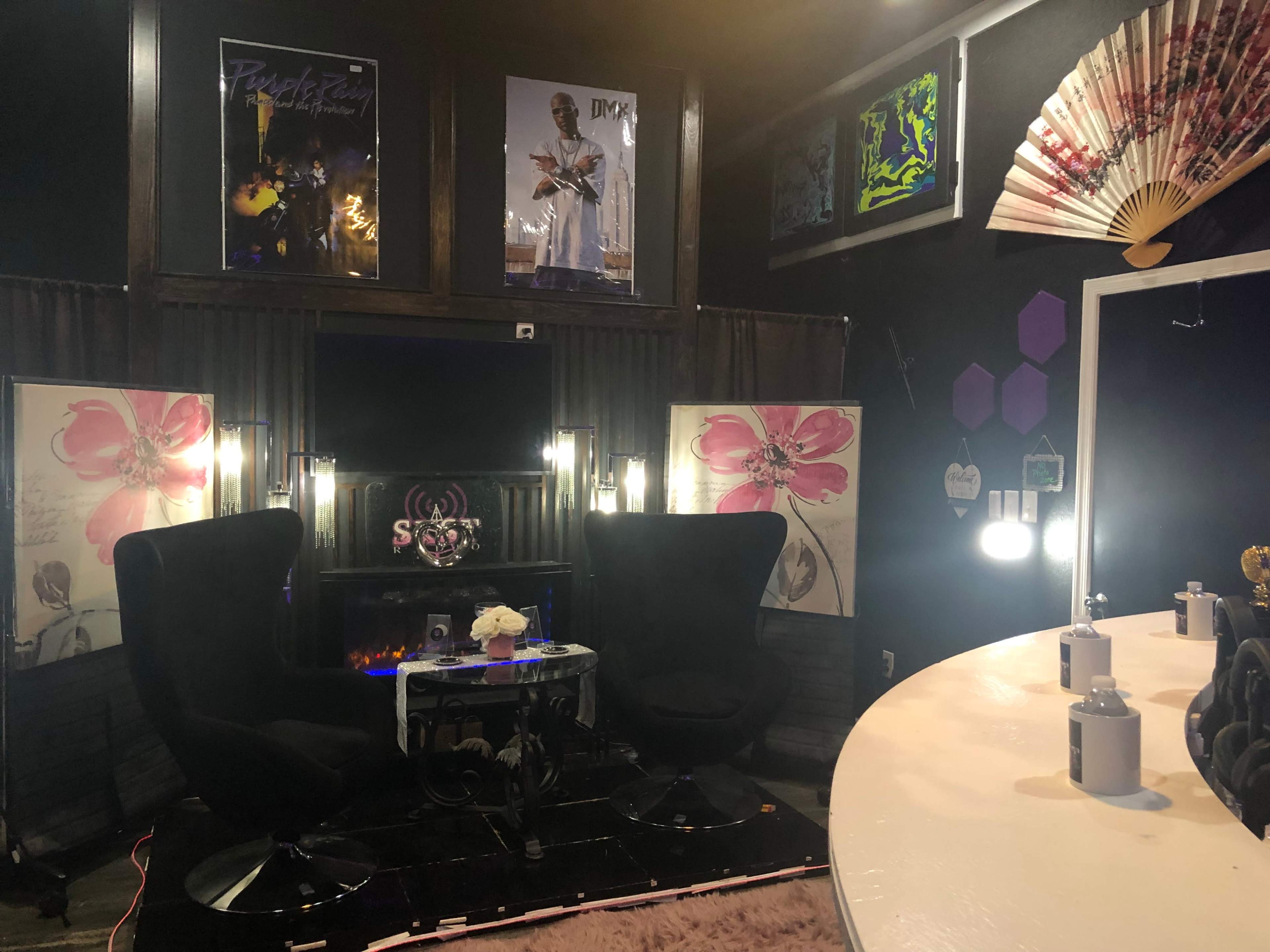 The image shows a stylish indoor space with two black chairs facing a small glass table, surrounded by decorative walls featuring artwork and ambient lighting.