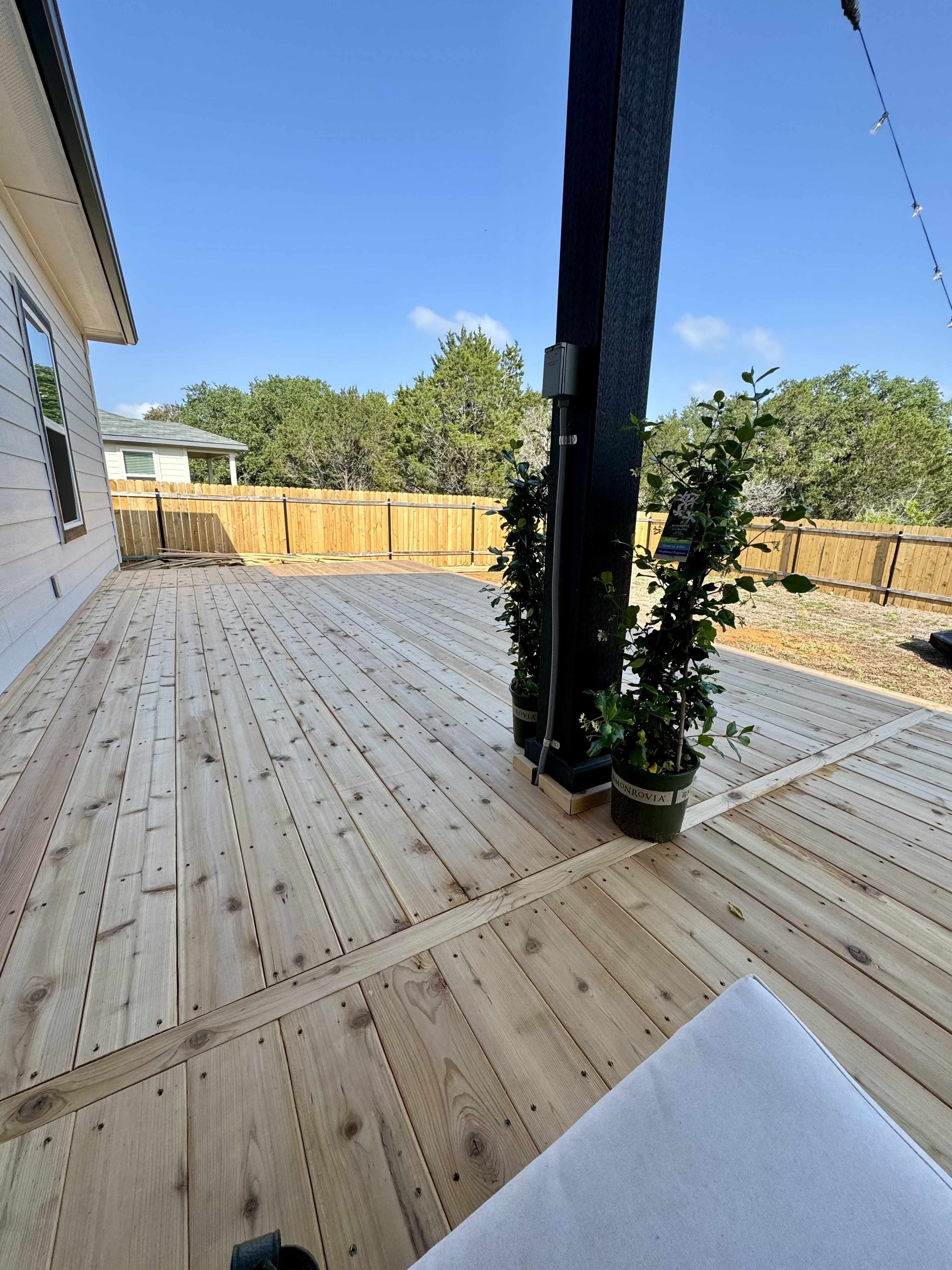 A wooden deck with planters featuring green plants, surrounded by a fenced yard and a clear blue sky.