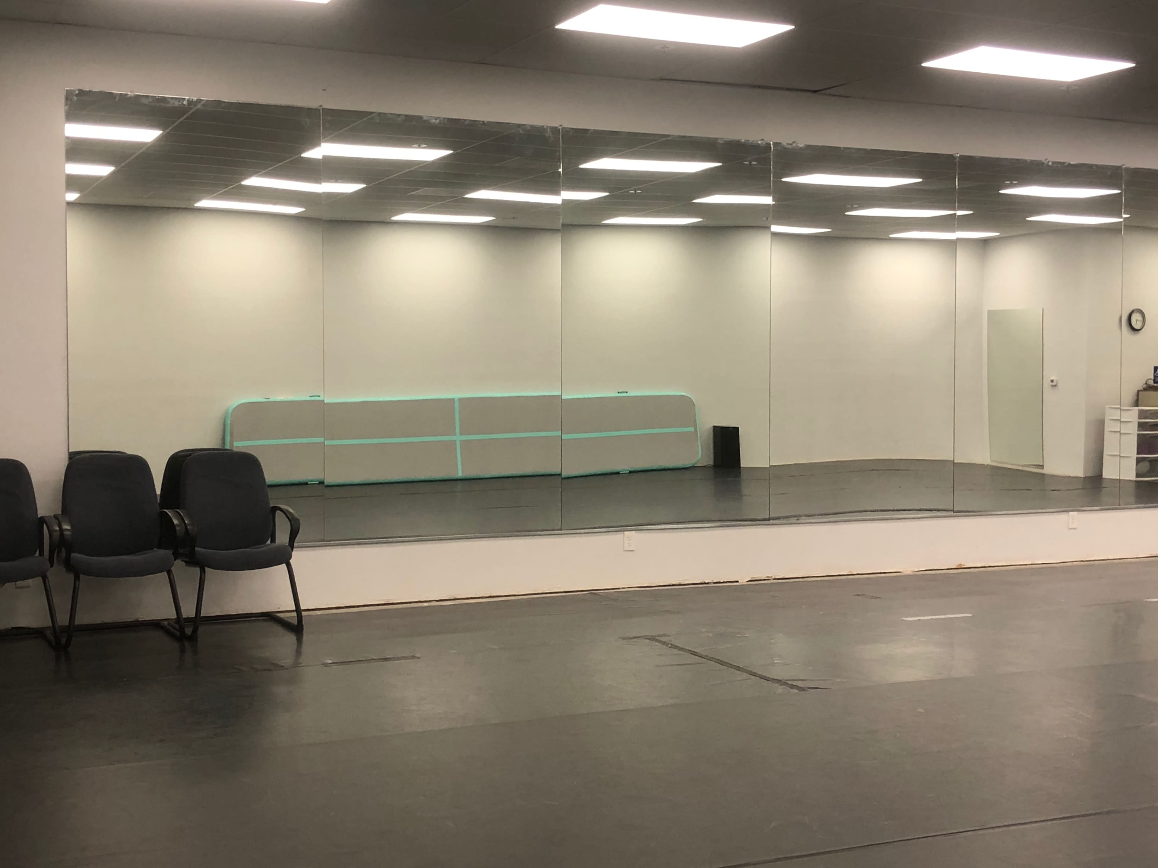 The image shows a dance studio with a mirrored wall, a barre, and empty chairs in front of a smooth black floor.