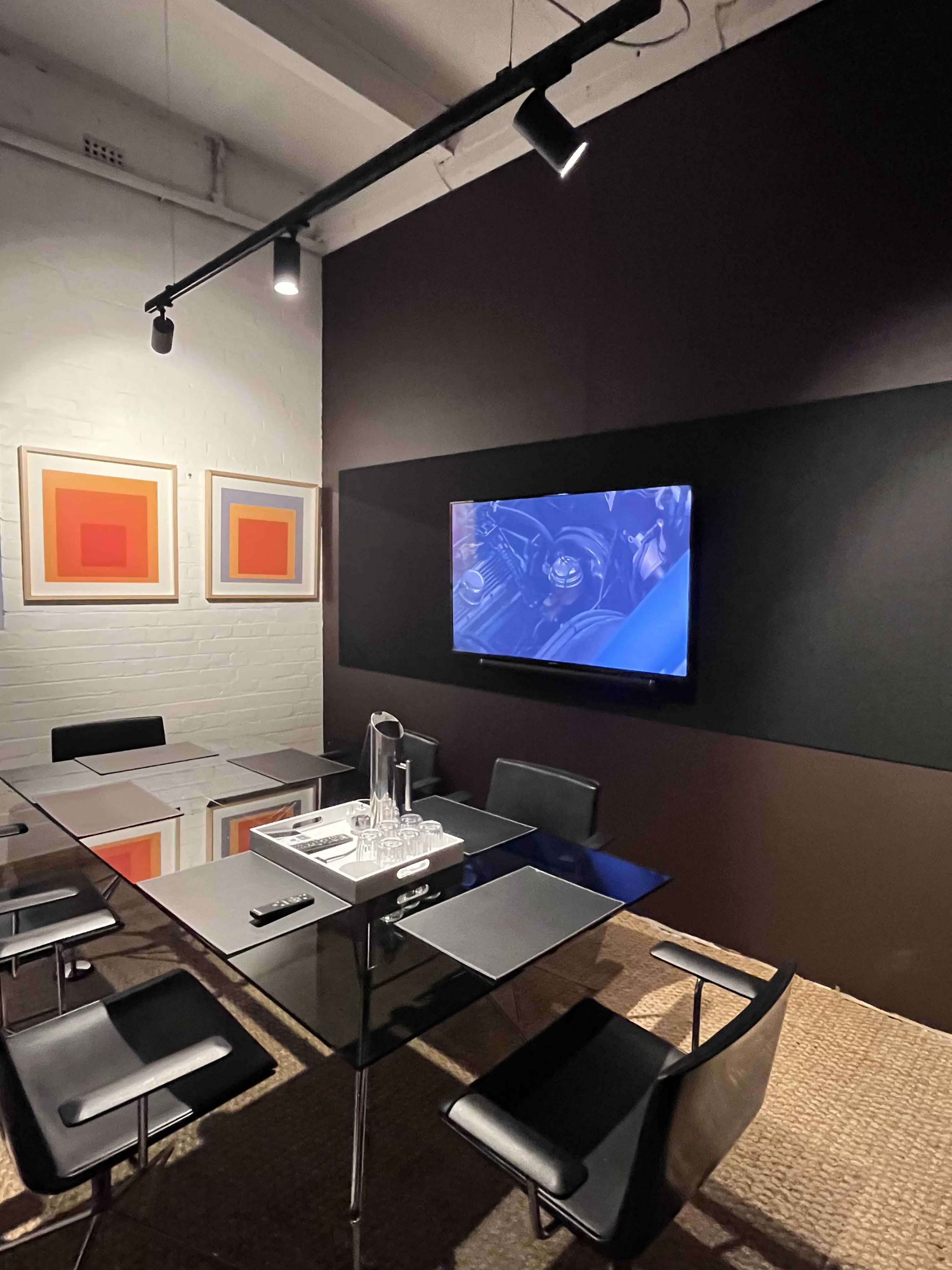 A conference room features a glass table surrounded by black chairs, with artwork on the walls and a television mounted on a dark panel.
