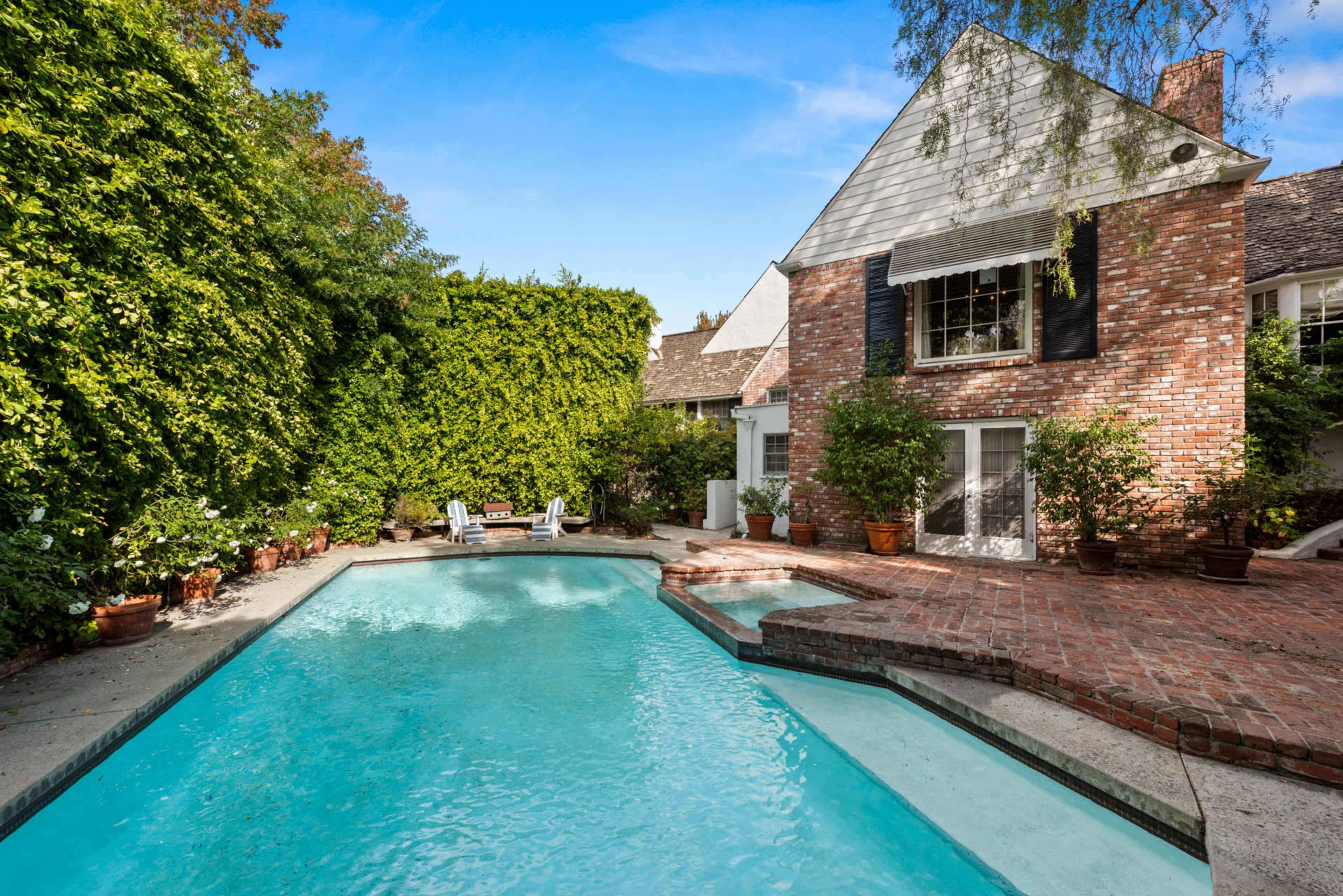 A brick house with white shutters is set beside a clear swimming pool surrounded by neatly arranged potted plants and a stone patio.