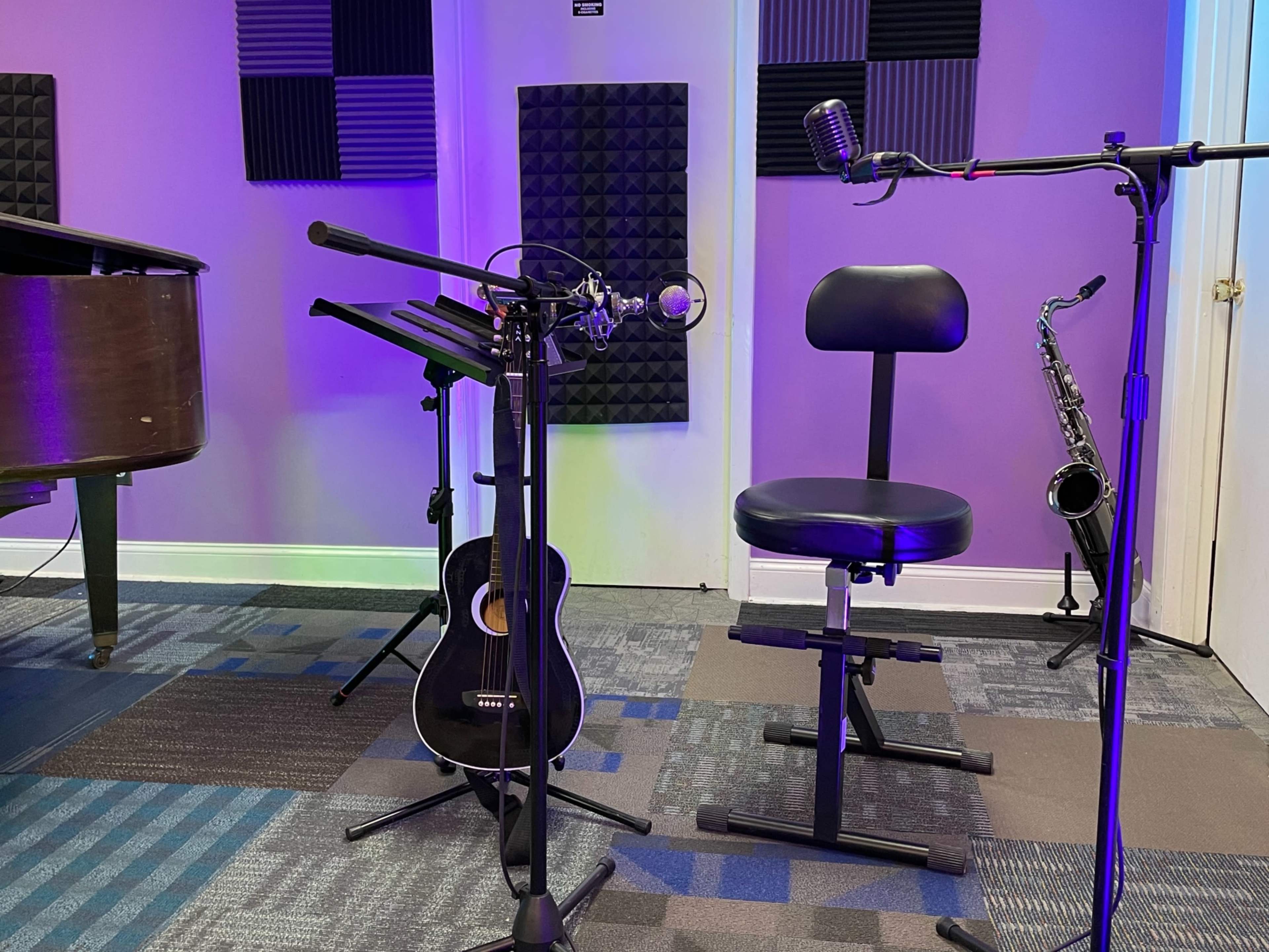 A microphone is positioned on a stand next to a black guitar and a saxophone, with a chair set in front and soundproof panels on the walls.