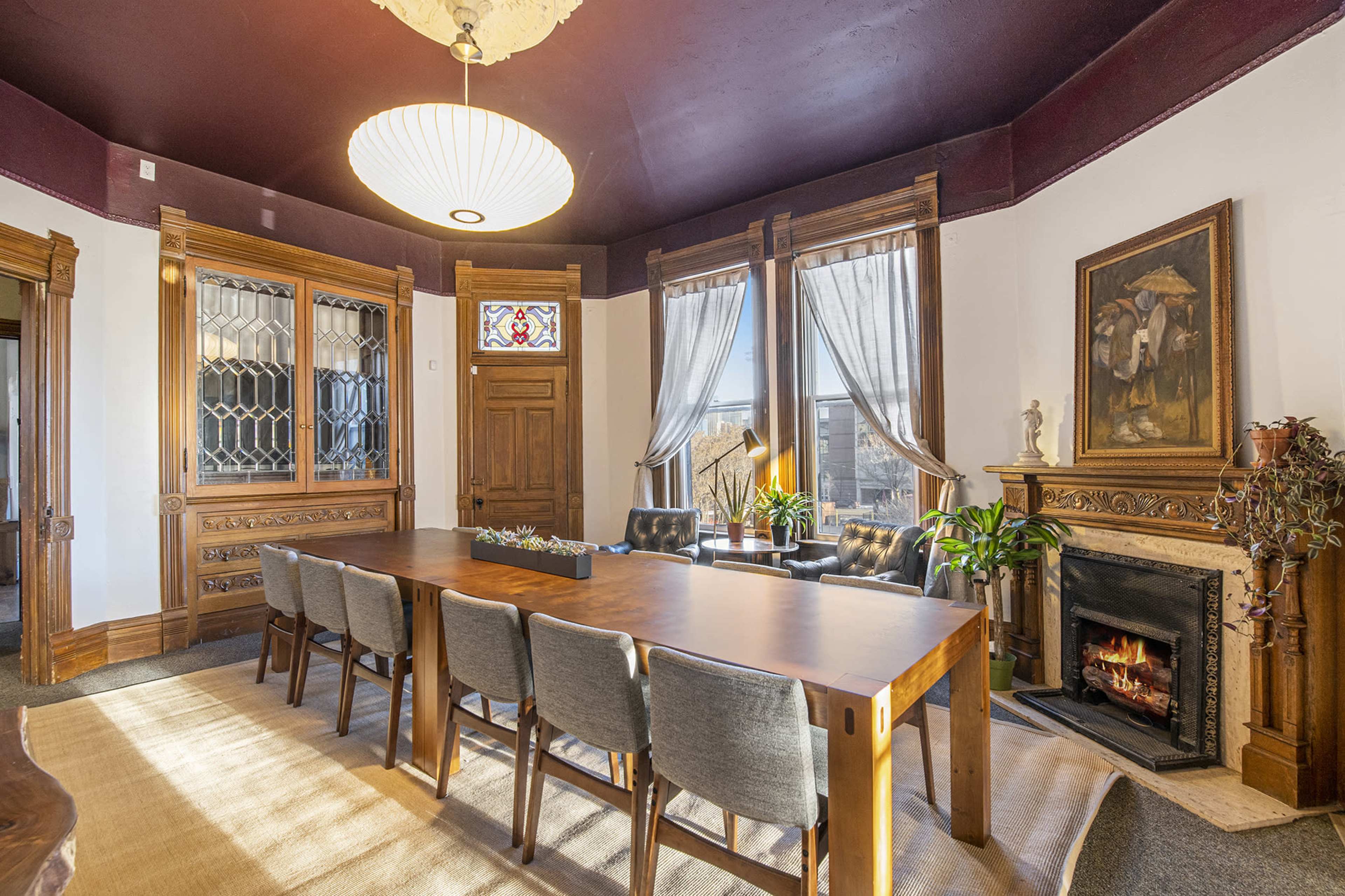 A spacious dining room featuring a large wooden table surrounded by chairs, with a fireplace and large windows adorned with curtains.