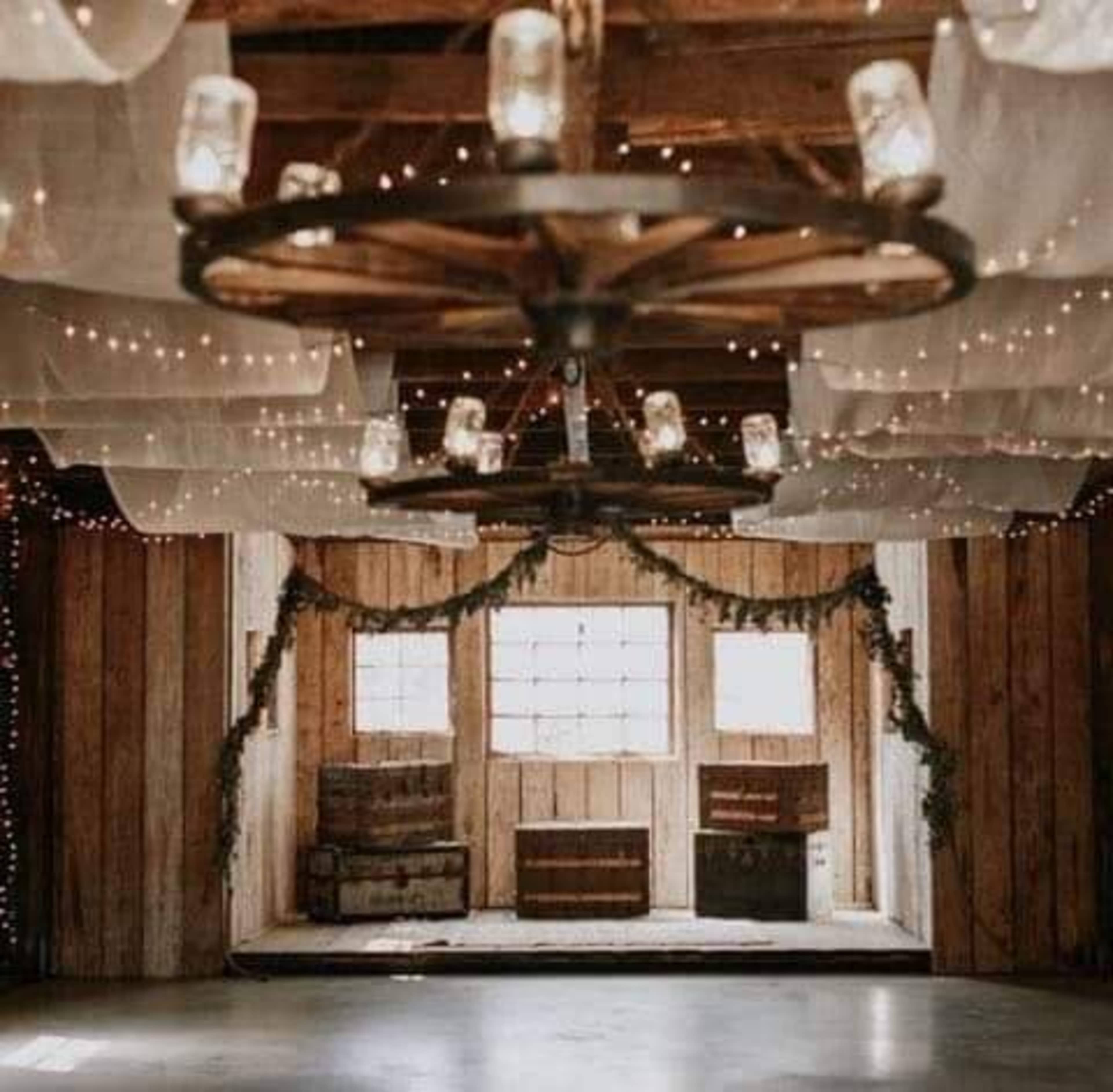 A rustic interior of a barn with wooden walls, a ceiling adorned with lights and draped fabric, and wooden crates positioned in front of large windows.