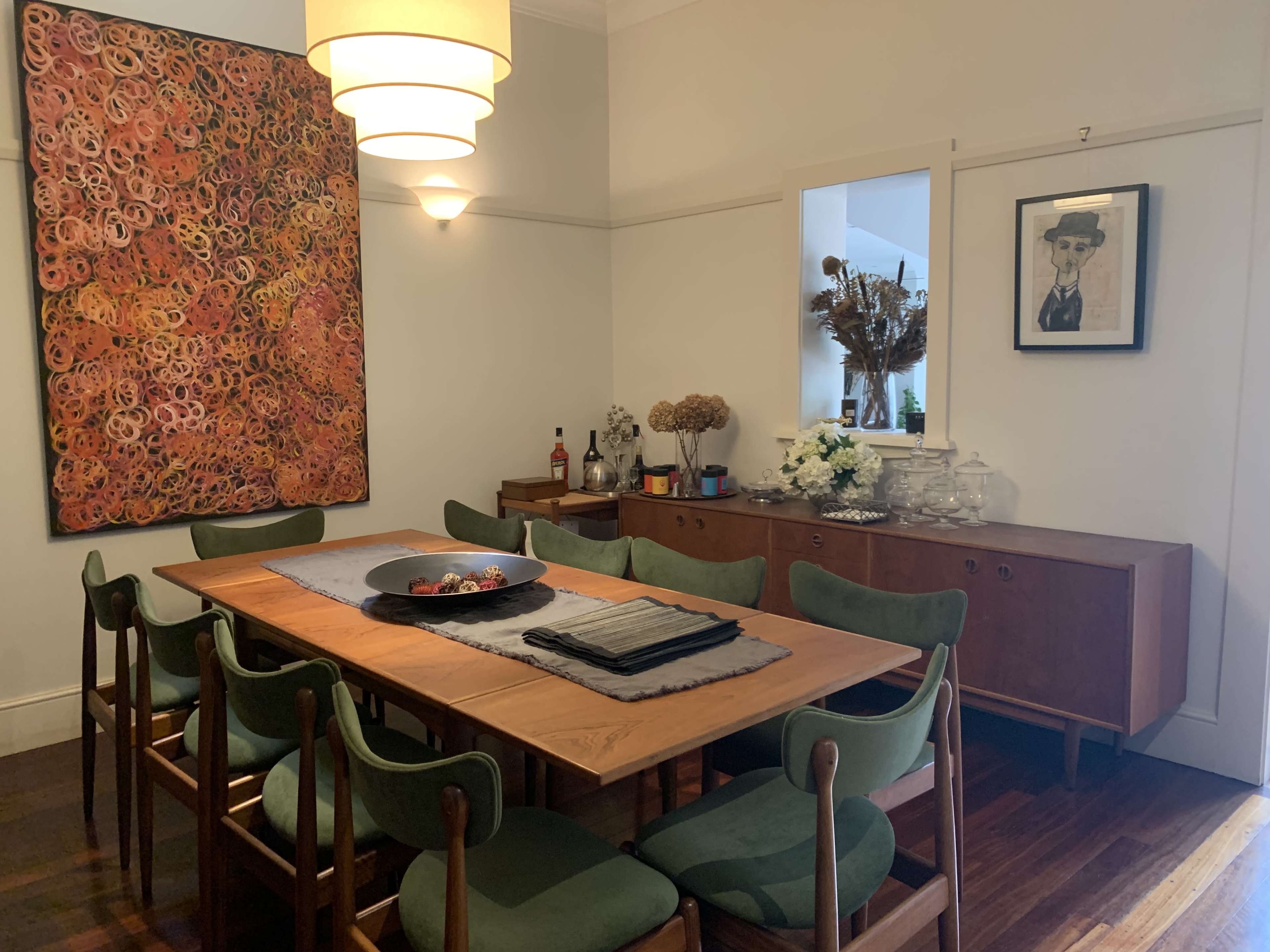 A dining room features a wooden table surrounded by green chairs, with a large abstract painting on the wall and a sideboard adorned with decorative items.