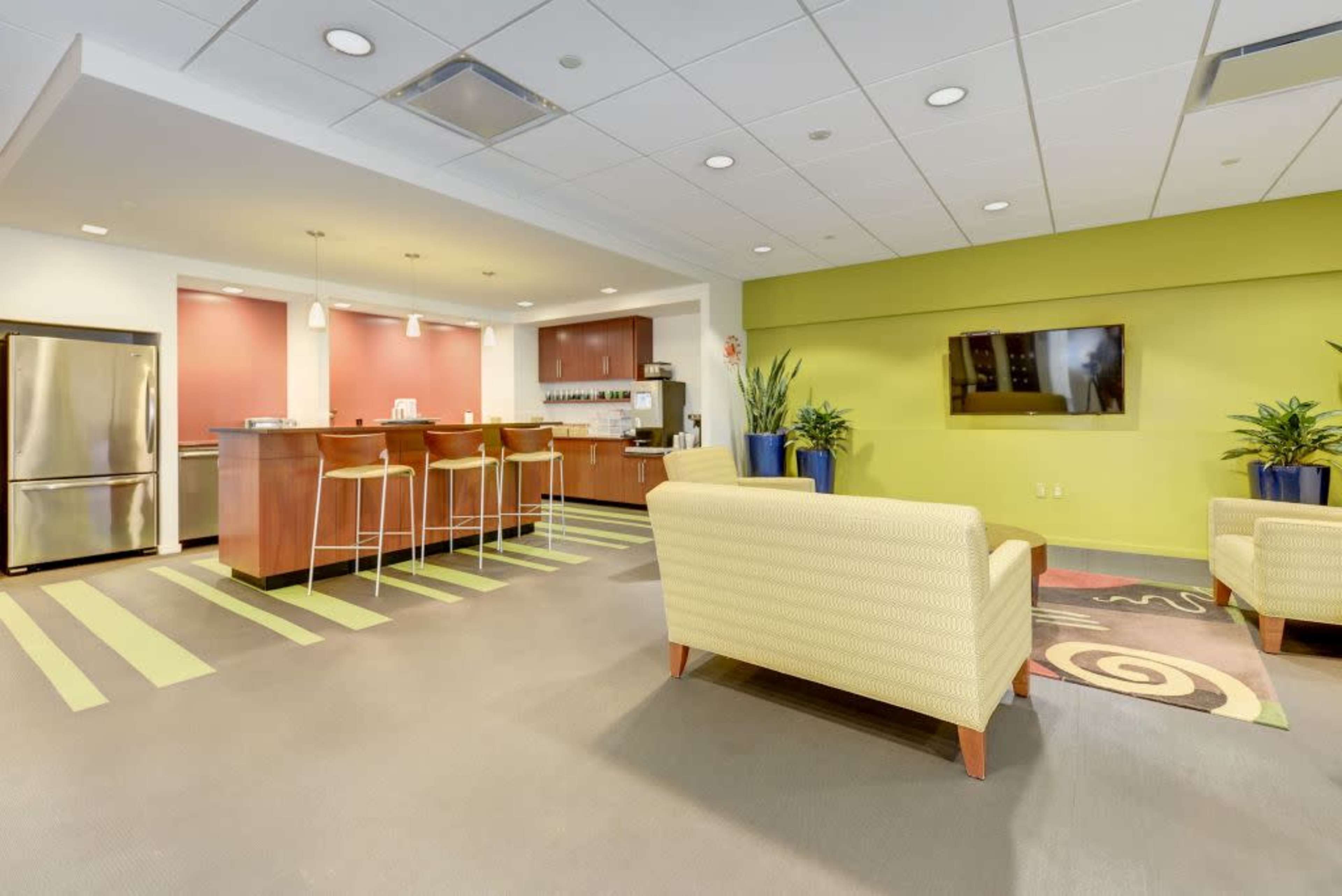 The image shows a modern communal lounge area with a kitchen, featuring bar stools, two armchairs, a television, and decorative plants.